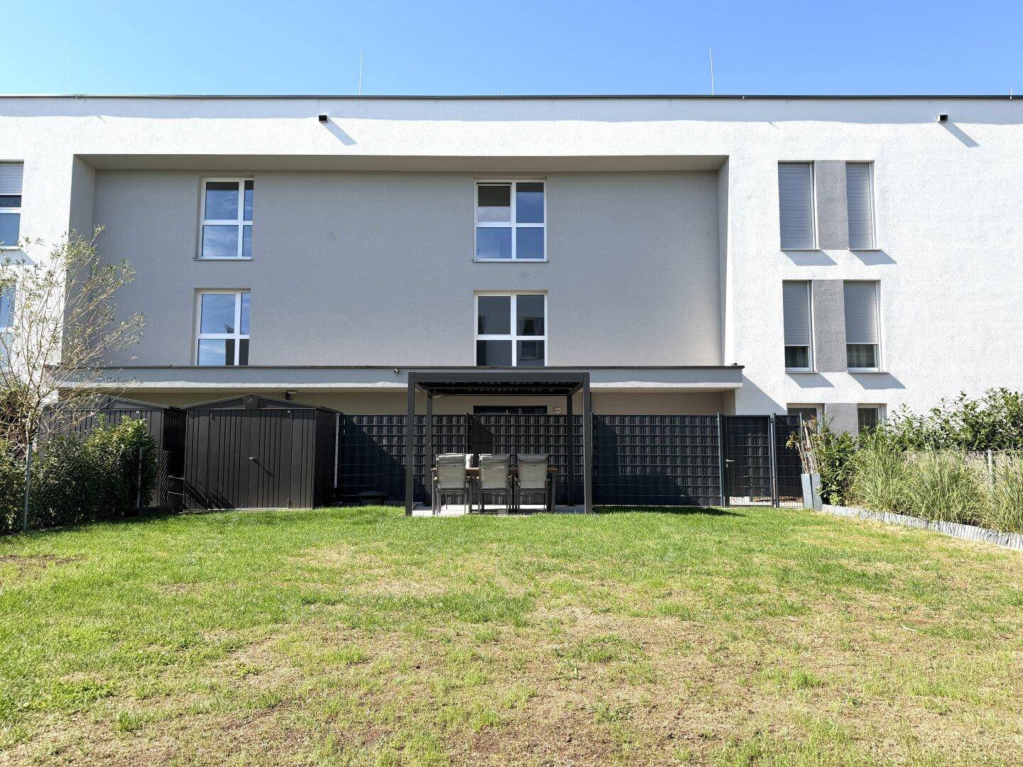Rückseite des Gebäudes mit gepflegtem Rasen, Gartenhaus und einem überdachten Sitzbereich.