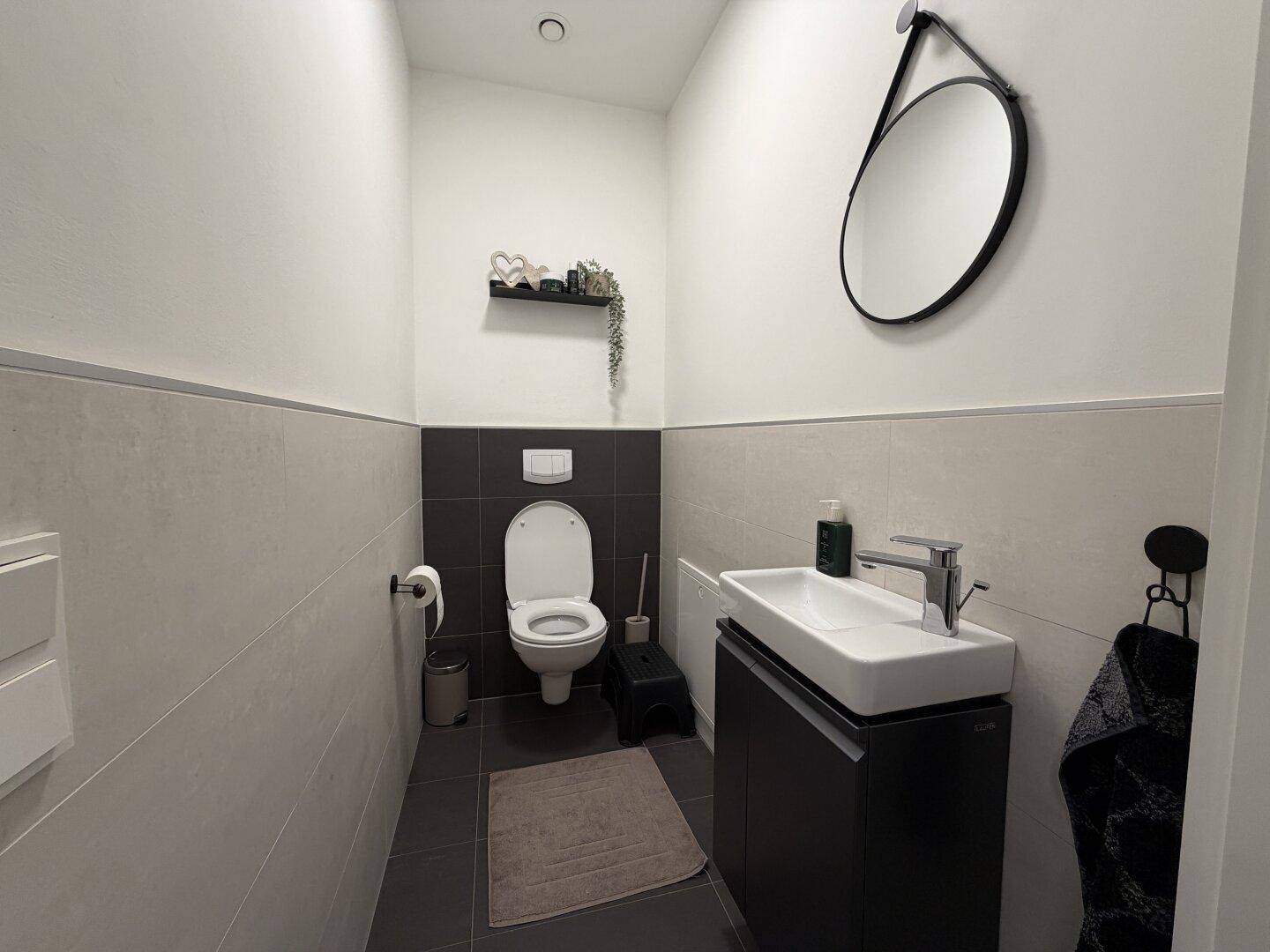 Modernes Gäste-WC mit Wand-WC, kleinem Waschbecken und stilvollem runden Spiegel.