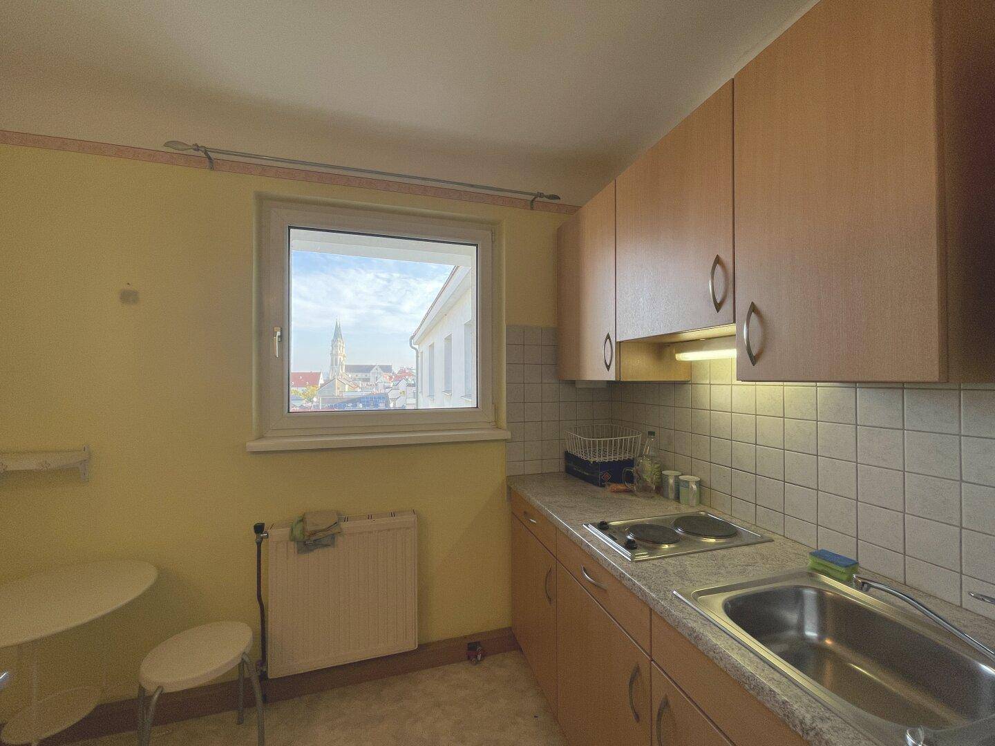 Helle Küche mit Holzschränken, Spüle, Kochfeld und einem Fenster mit Stadtblick.