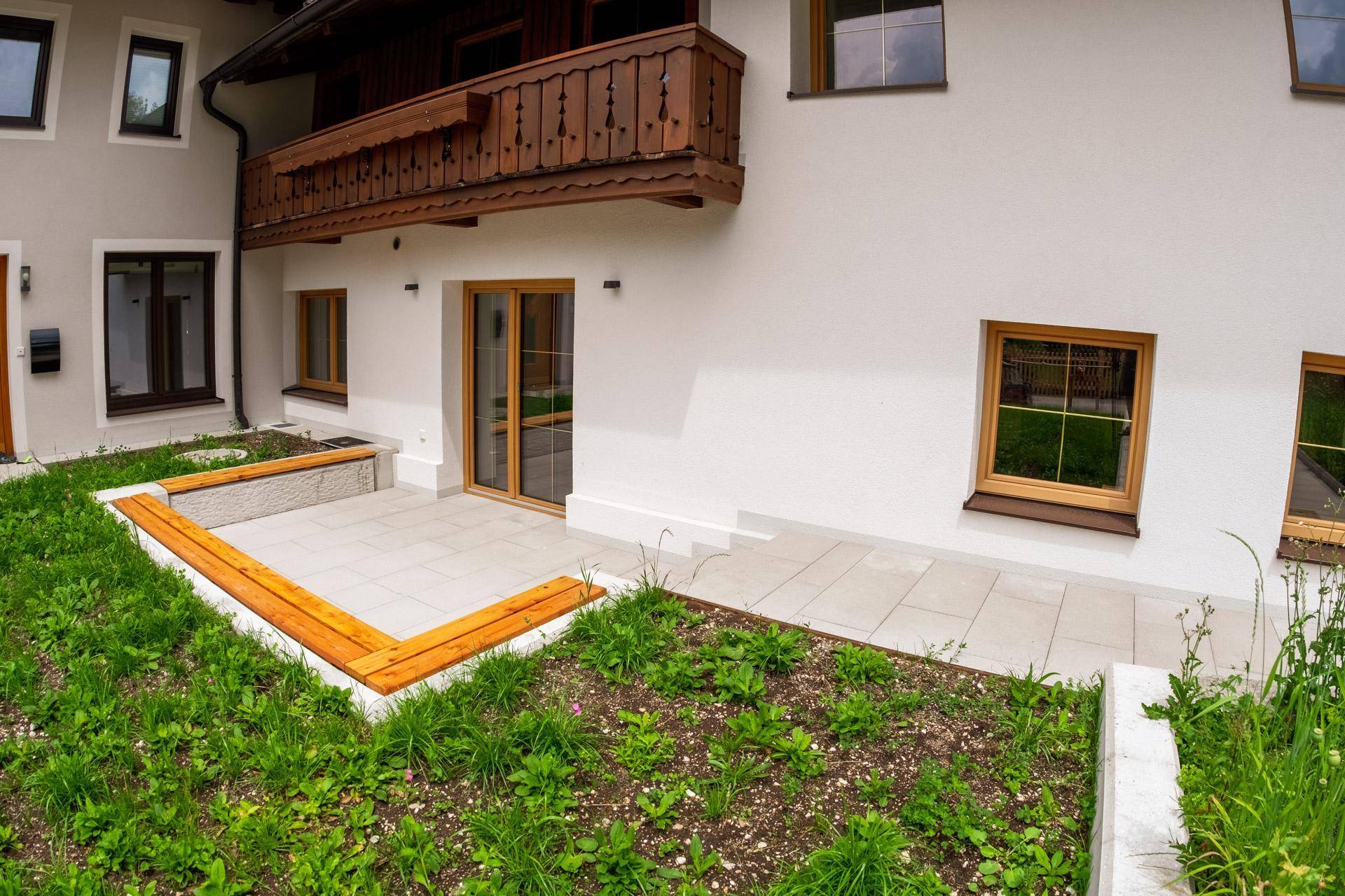 Helle, gepflasterte Terrasse mit Holzbänken und Zugang zum Gartenbereich eines Neubaus.