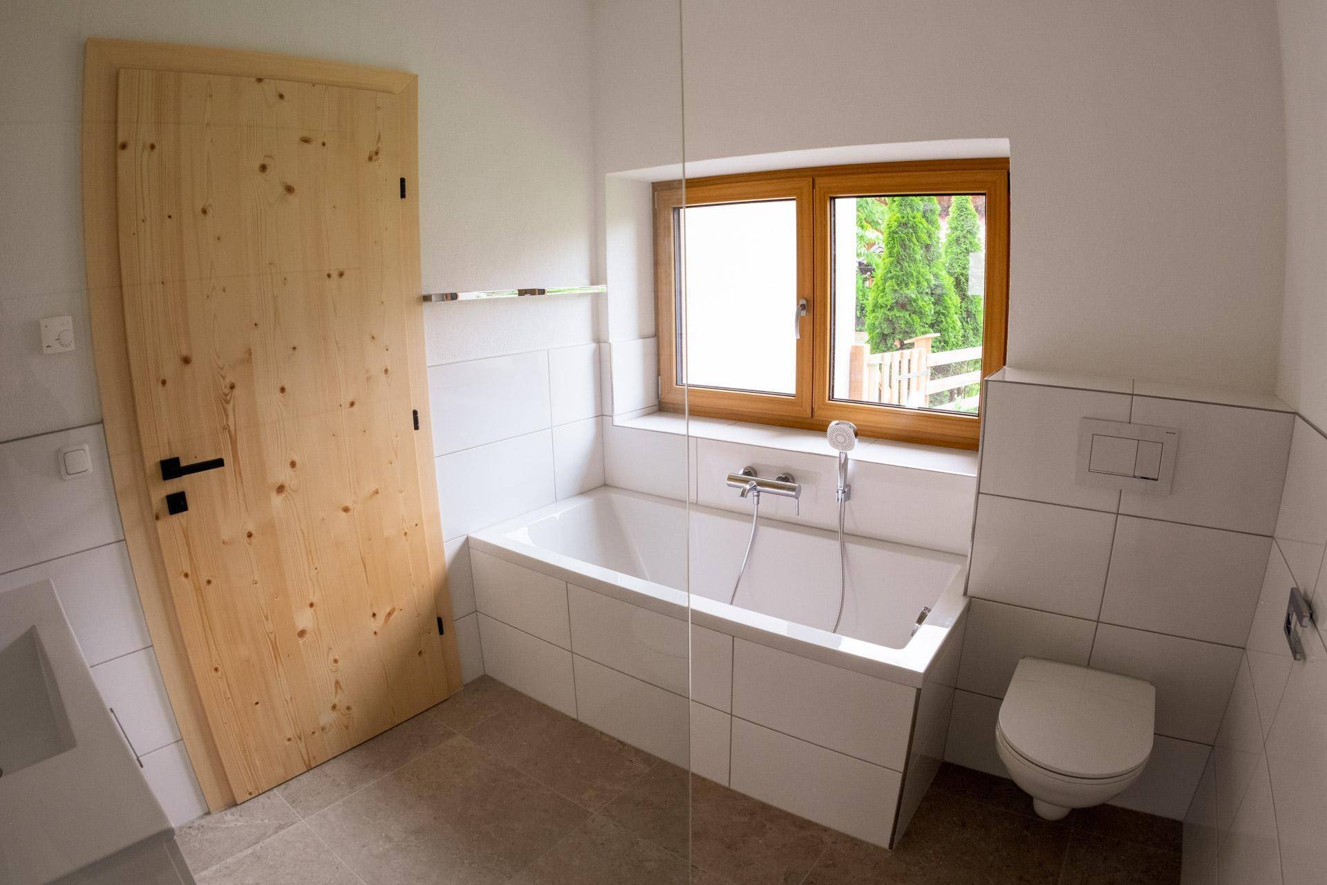 Helles Badezimmer mit Badewanne, Toilette, Fenster und weißen Wandfliesen.