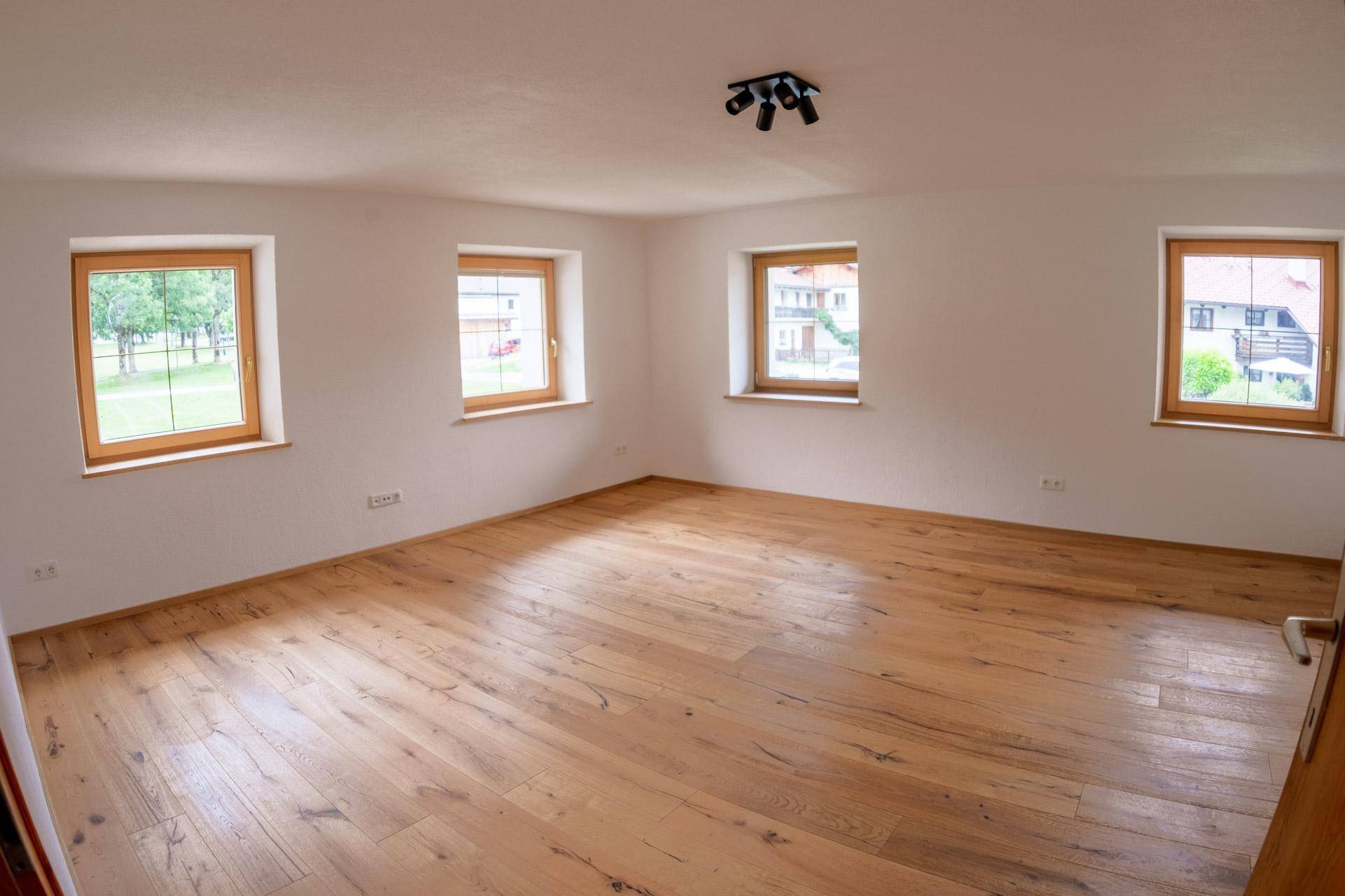 Großer, heller Wohnraum mit Holzboden, weißen Wänden und mehreren Fenstern.