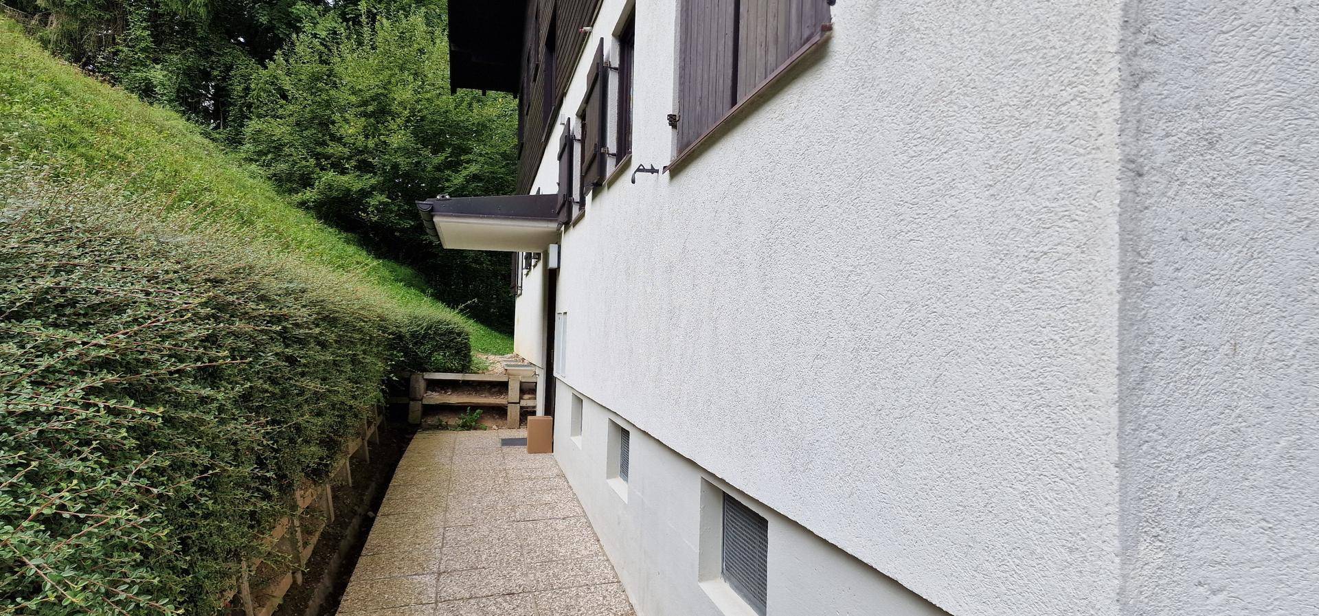 Weiße Hausfassade mit gepflastertem Weg und grüner Hecke, die den Hang hinaufführt.