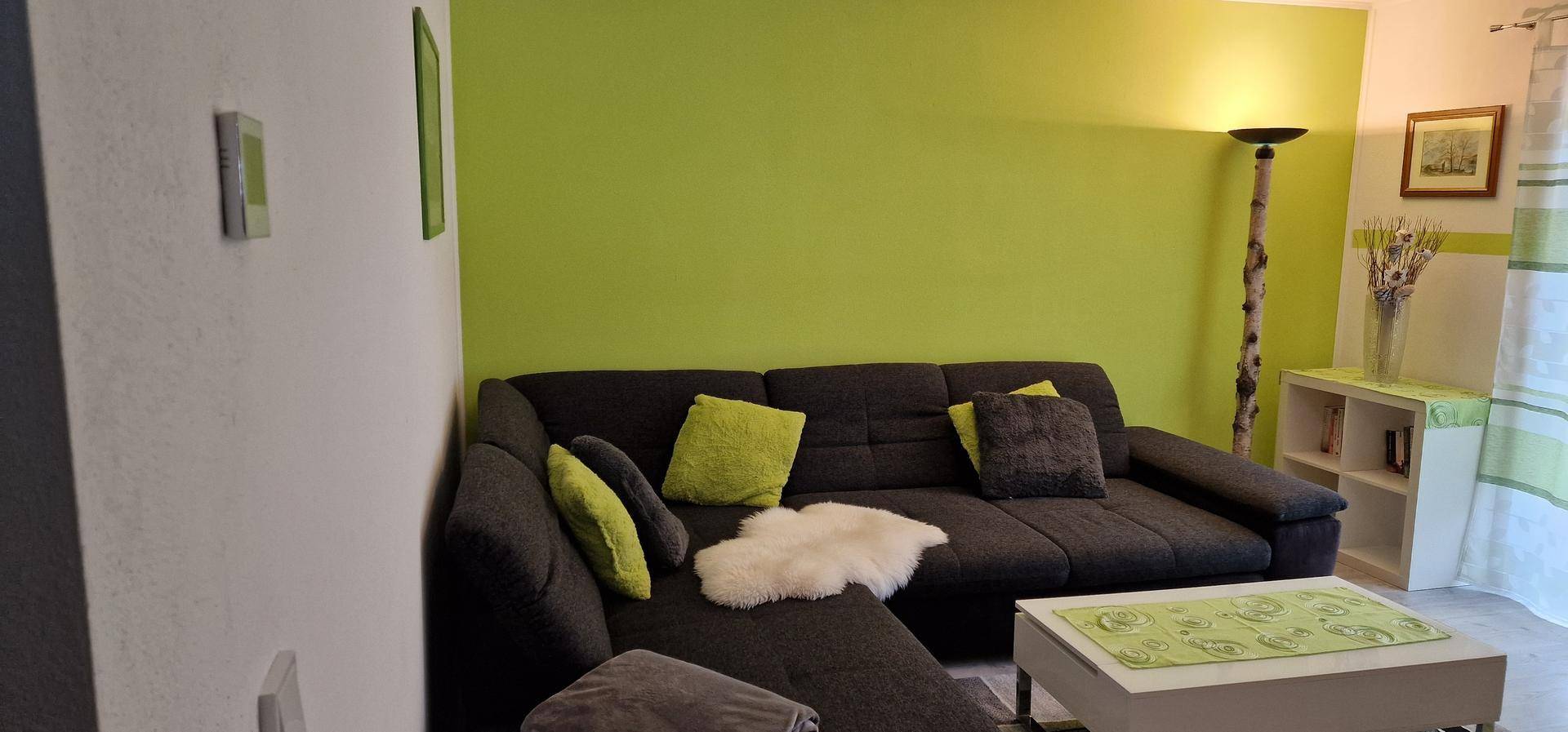 Helles Wohnzimmer mit grüner Akzentwand, dunklem Ecksofa und modernem Stehlampen-Design.