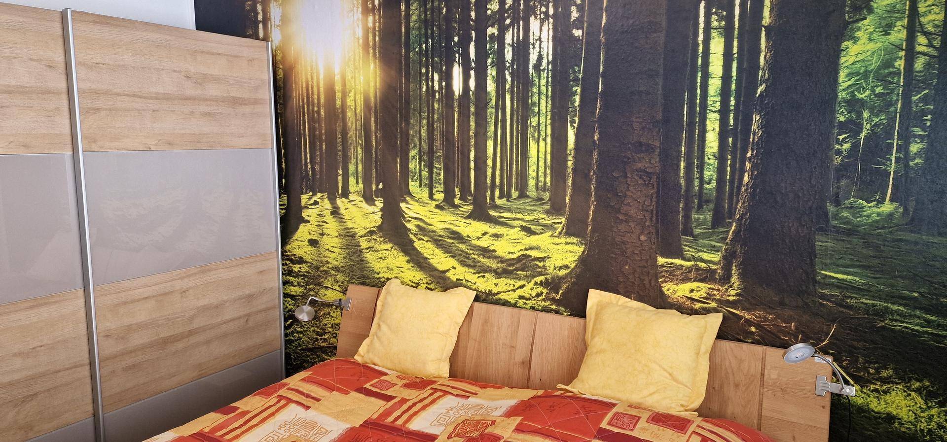 Schlafzimmer mit Waldmotiv-Tapete hinter dem Bett und hellem Kleiderschrank, der viel Stauraum bietet.