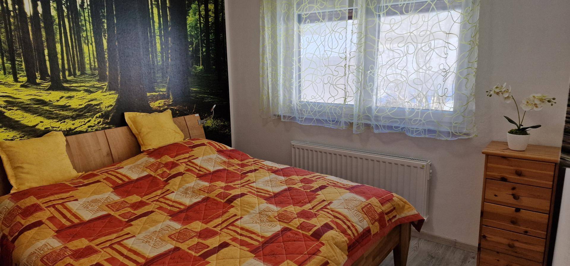 Schlafzimmer mit Doppelbett, Waldmotiv-Tapete und hellem Fenster, das viel Tageslicht hereinlässt.