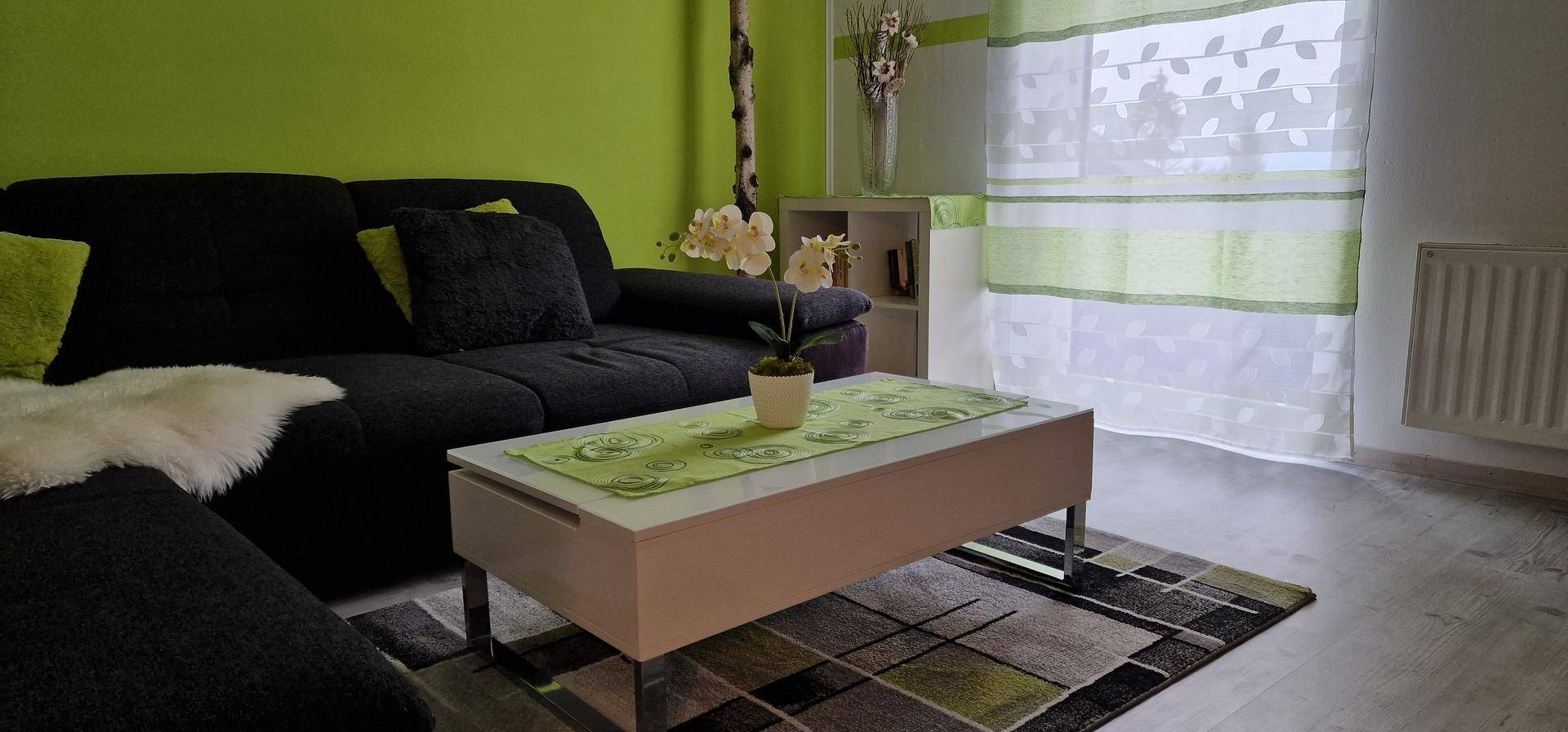 Gemütliches Wohnzimmer mit dunklem Sofa, Couchtisch und hellem Fenster mit grünen Vorhängen.