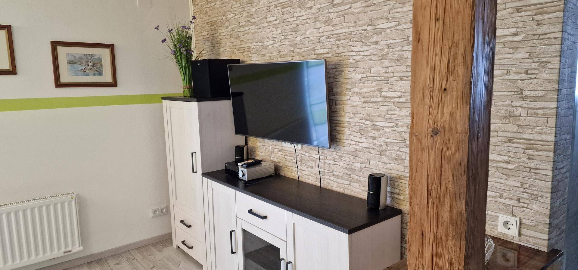 Wohnbereich mit Steinwand-Optik, Flachbildfernseher und rustikalem Holzbalken als Raumteiler.