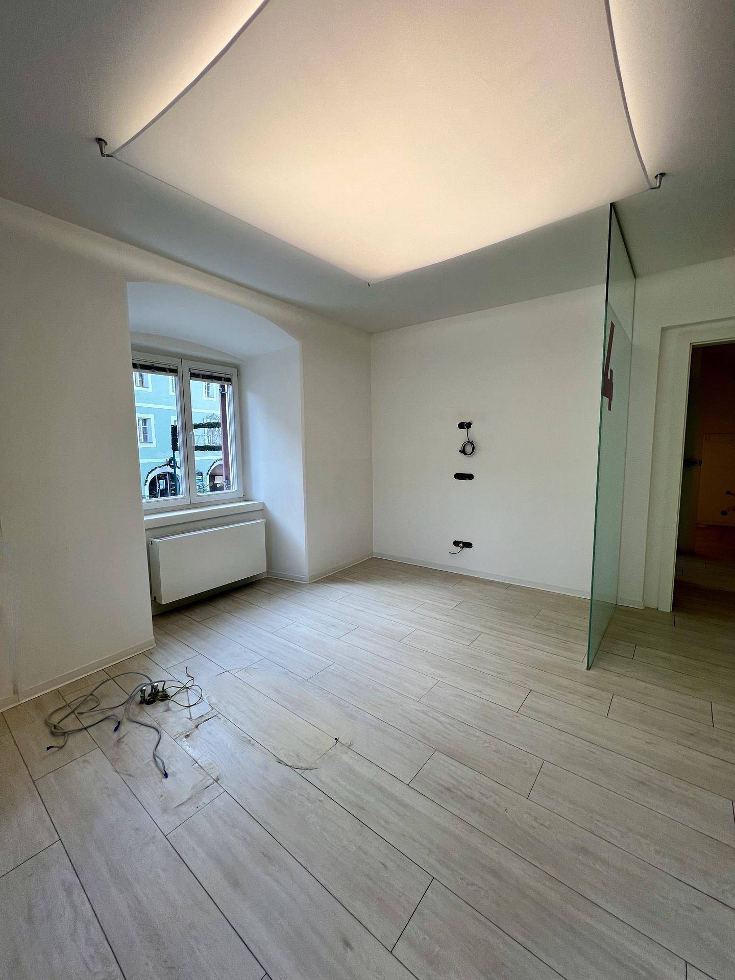 Leerer Büroraum mit weiß gestrichenen Wänden, hellem Fliesenboden und einem Fenster mit Blick nach draußen.