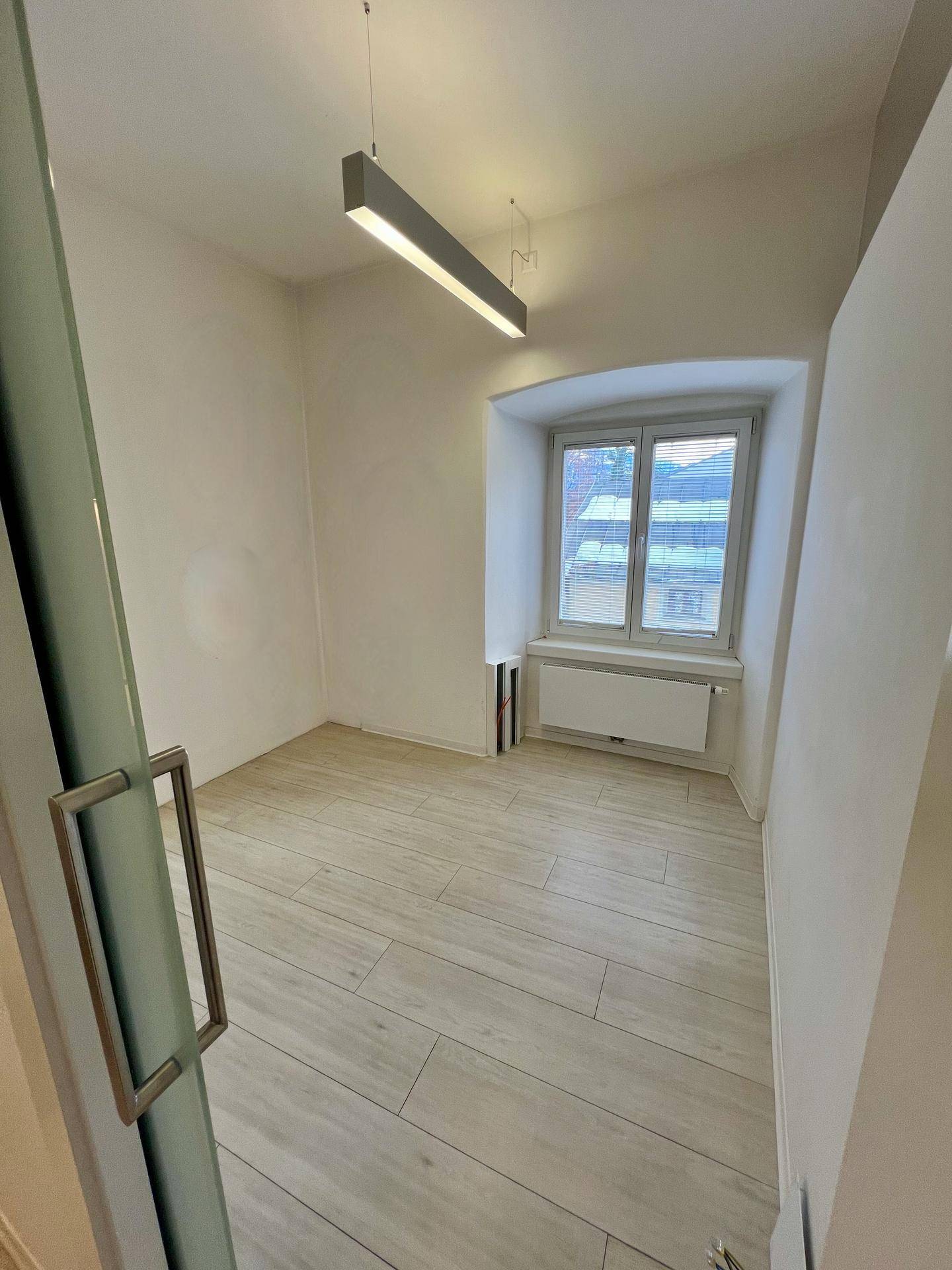 Kleiner, heller Büroraum mit einem Fenster, das viel Tageslicht hereinlässt, und einem Heizkörper.