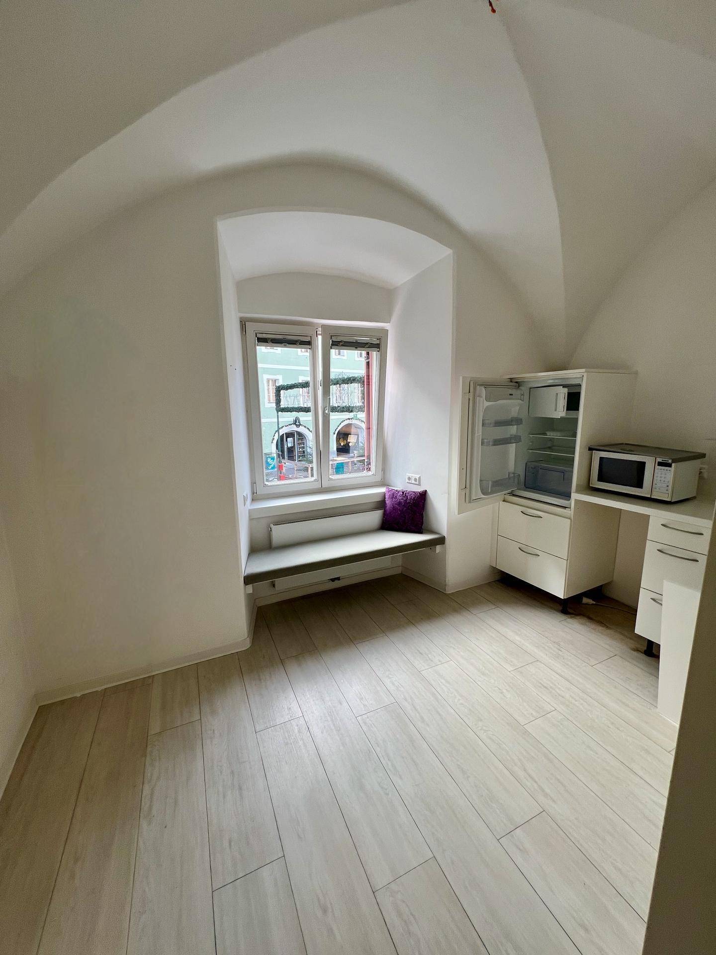 Kleiner Raum mit Fenster, integrierter Sitzbank, Minikühlschrank und Mikrowelle, unter Gewölbedecken.
