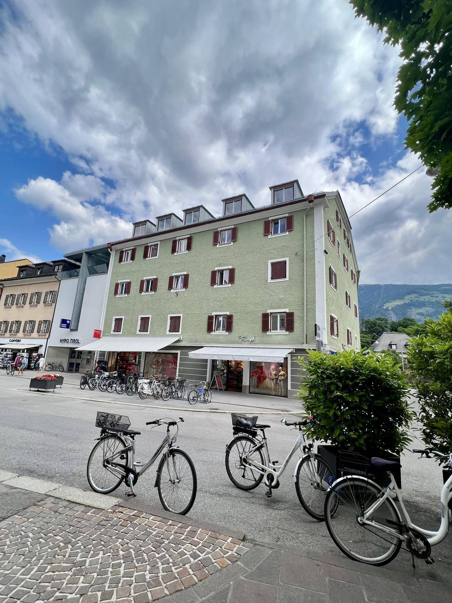 Grünes Gebäude mit vielen Fenstern und Geschäften im Erdgeschoss an einer belebten Straße mit Fahrrädern.