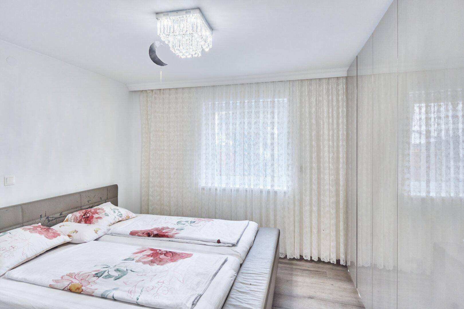 Schlafzimmer mit Doppelbett, weißem Hochglanz-Kleiderschrank und großen Fenstern für viel Tageslicht.