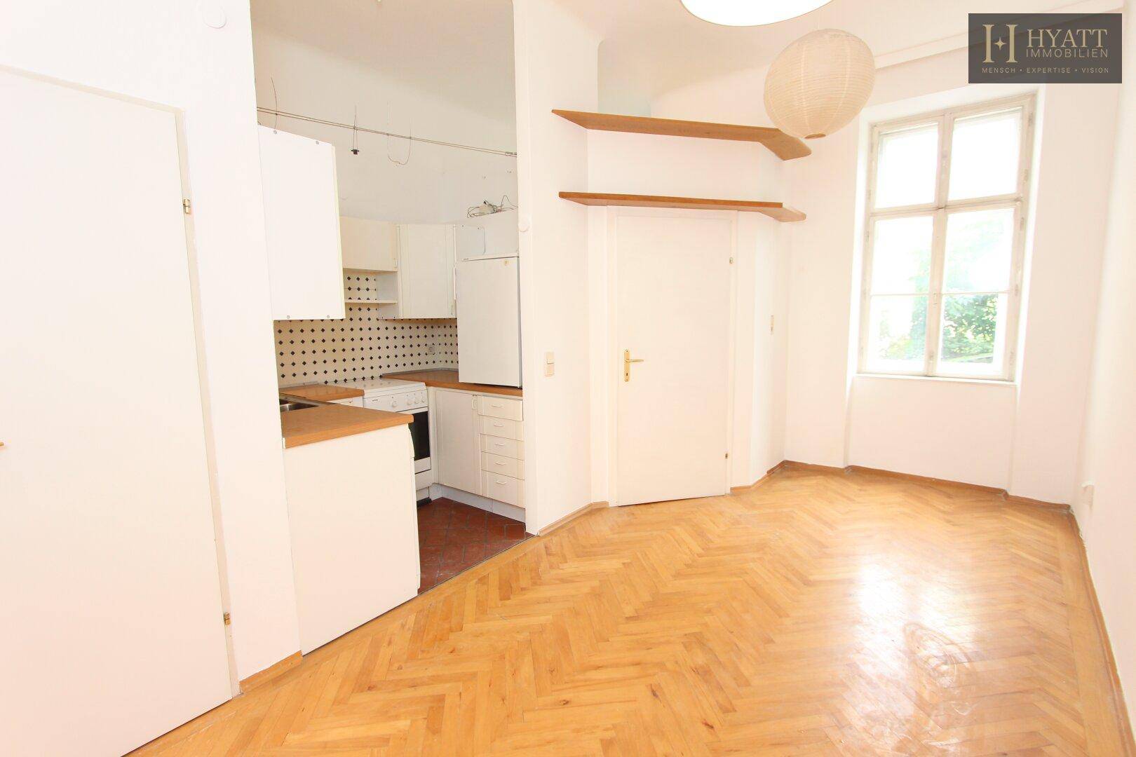 Heller Wohnraum mit Fischgrätparkett, integrierter Küchenzeile und großem Fenster.