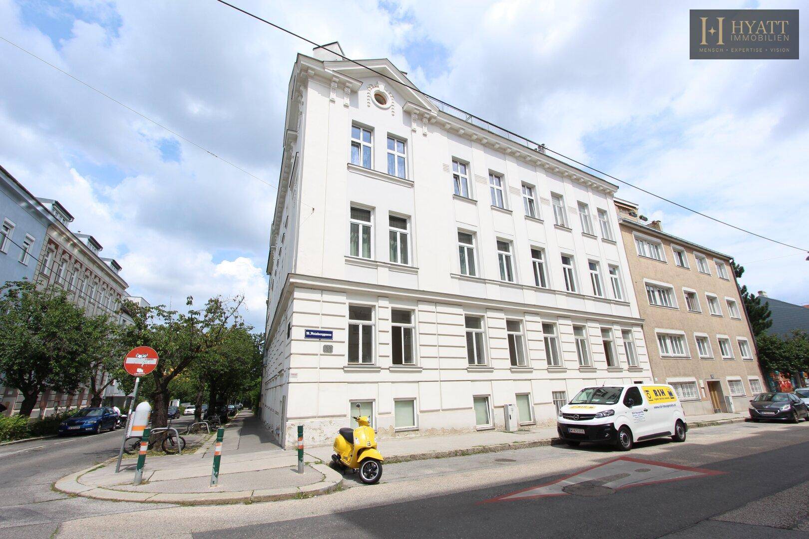 Weißes Eckgebäude mit klassischer Architektur und vielen Fenstern an einer städtischen Straße.