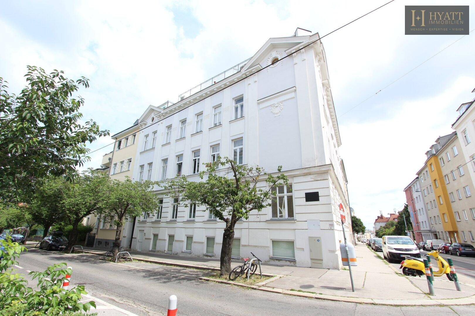 Historisches Eckgebäude mit weißer Fassade und Bäumen entlang der Straße.