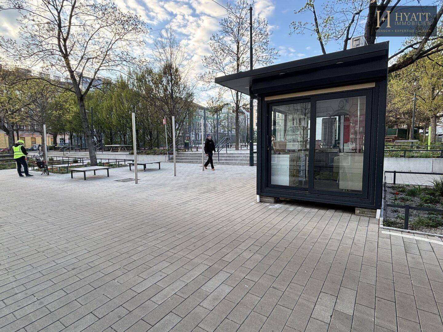 Ansicht des Kiosks auf dem öffentlichen Platz mit Passanten und Bänken im Hintergrund.