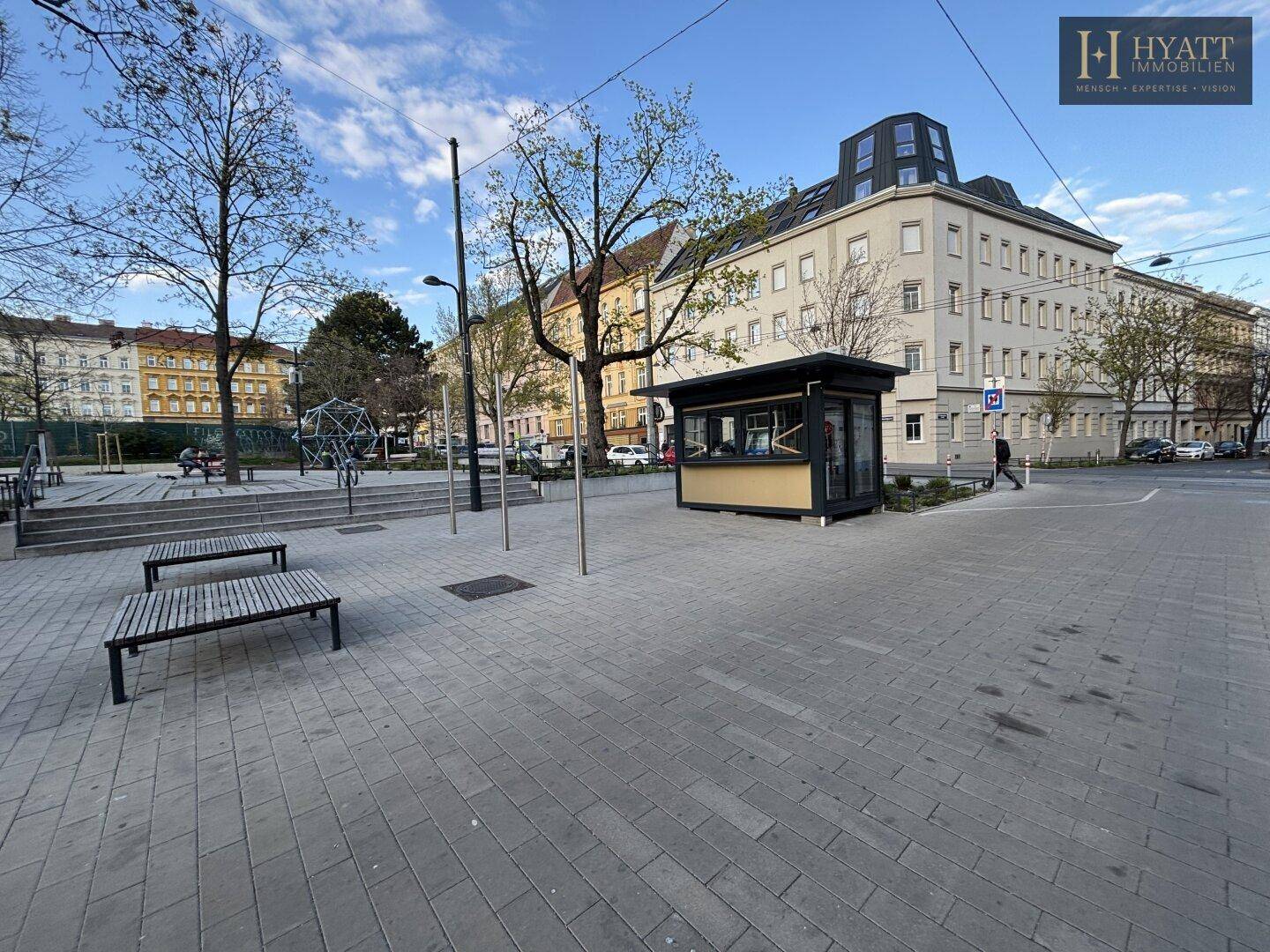 Weitwinkelaufnahme des Kiosks auf dem belebten Platz mit Bäumen und Sitzgelegenheiten.