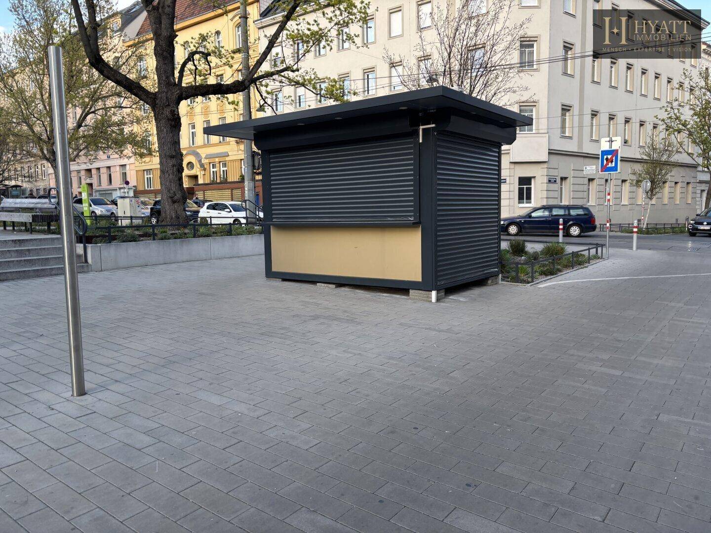 Seitenansicht des geschlossenen Kiosks mit Rollläden und einem gepflasterten Bereich davor.
