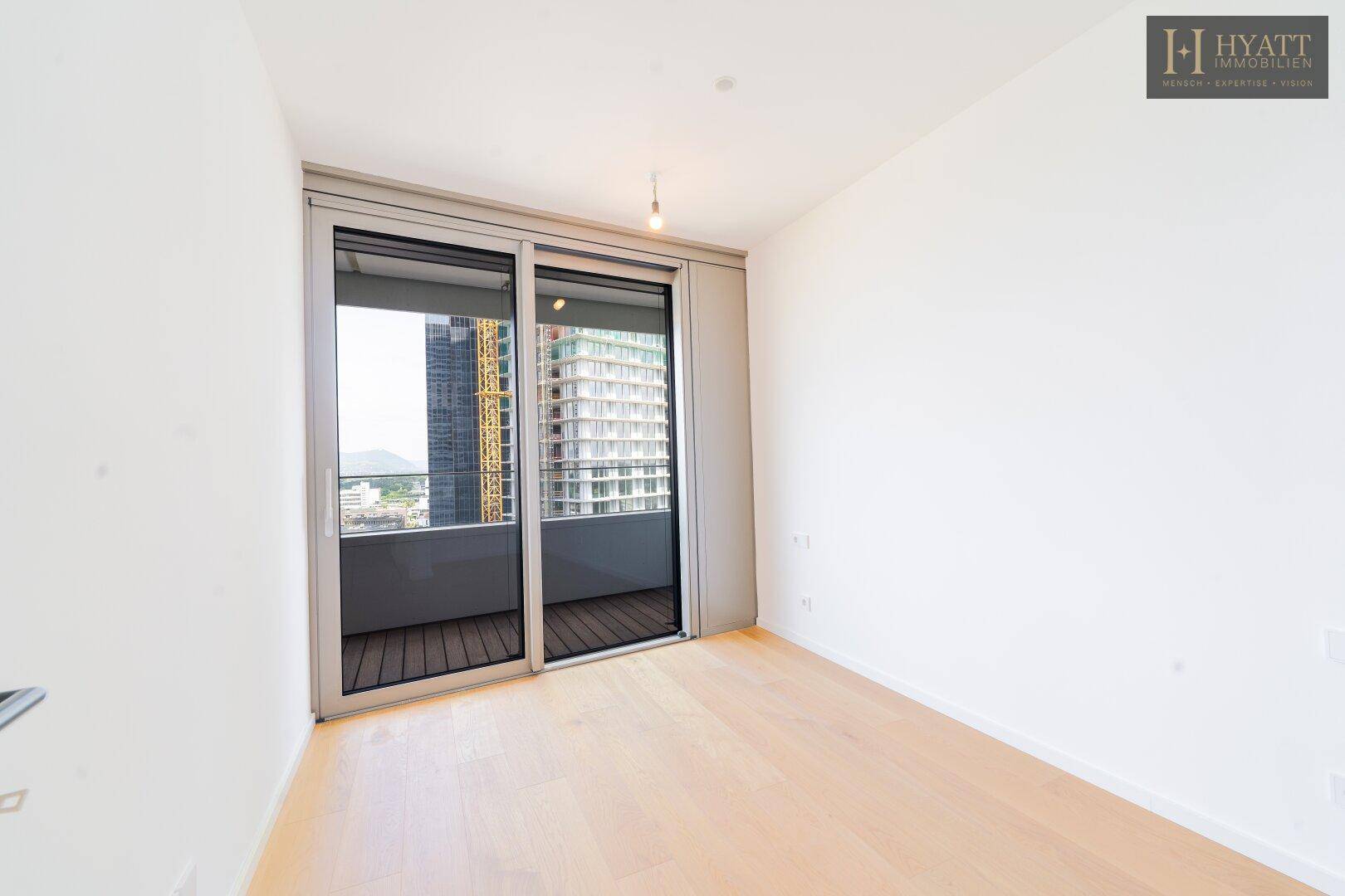 Helles Schlafzimmer mit Holzboden und Schiebetür zum Balkon, mit Blick auf die umliegenden Gebäude.