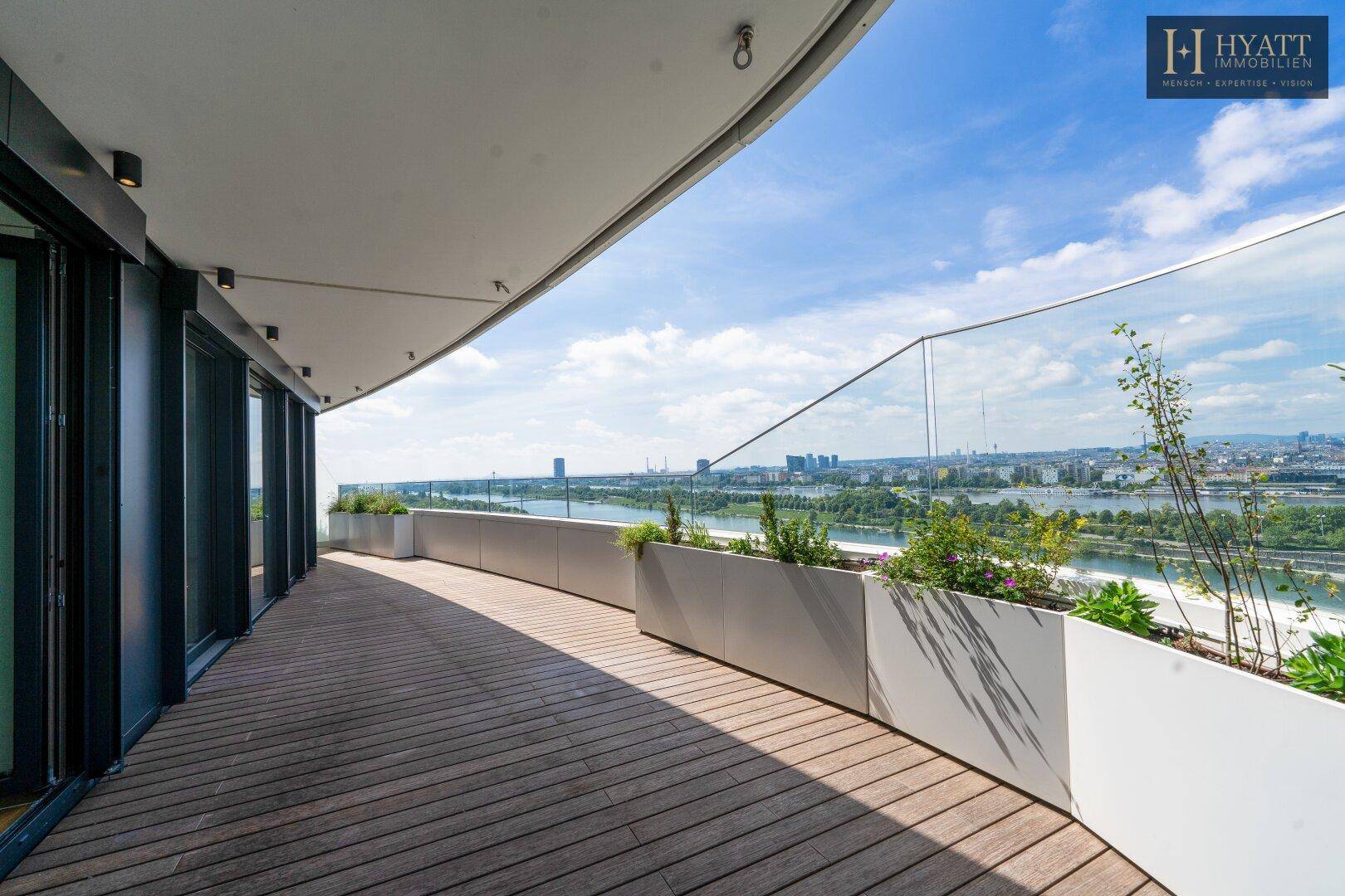 Geräumiger Balkon mit Holzboden und Pflanzkübeln, der einen weiten Blick über die Stadt und den Fluss bietet.