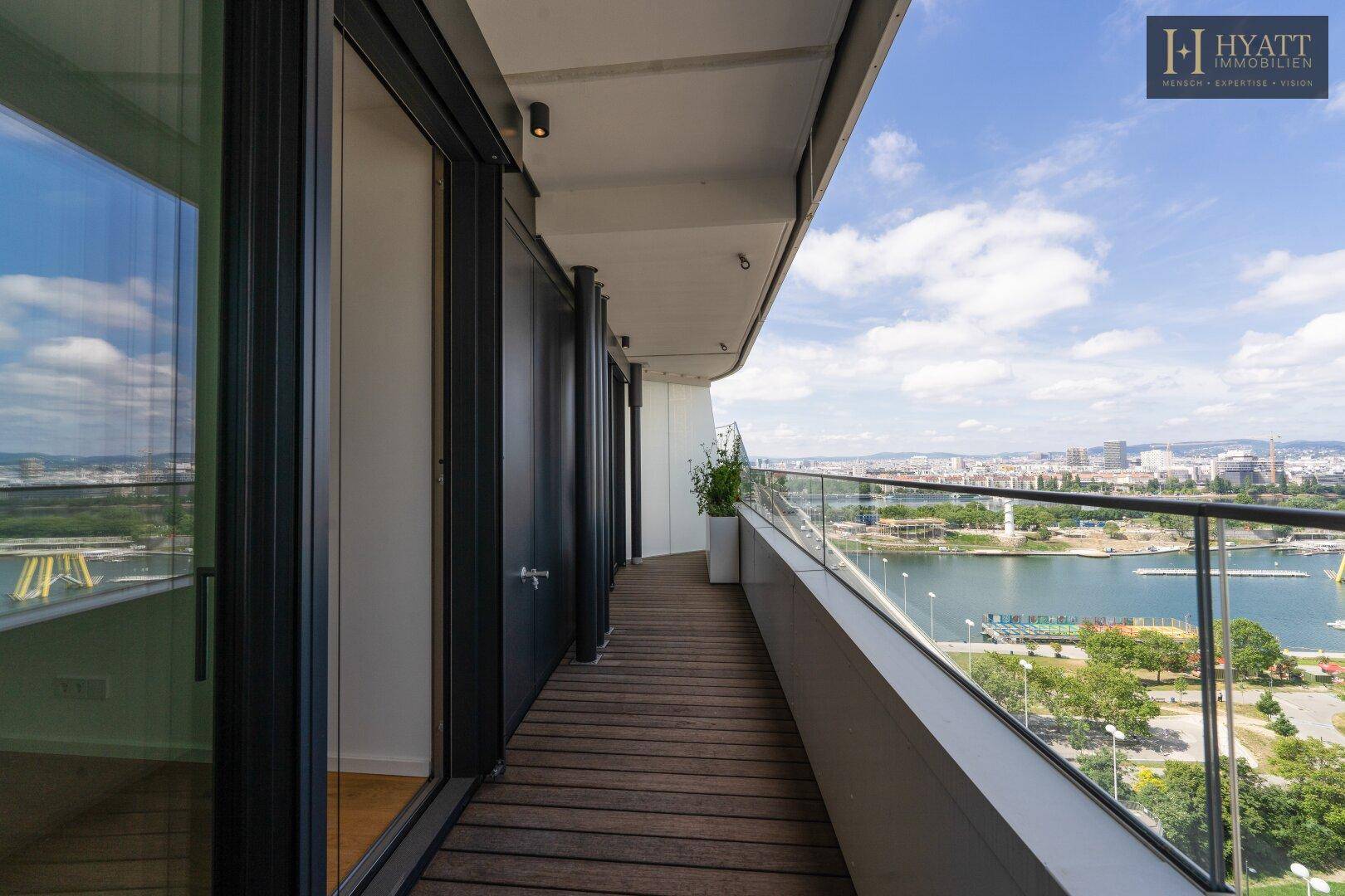 Schmaler Balkon mit Holzboden und Glasgeländer, der einen Panoramablick auf die Stadt und das Wasser bietet.