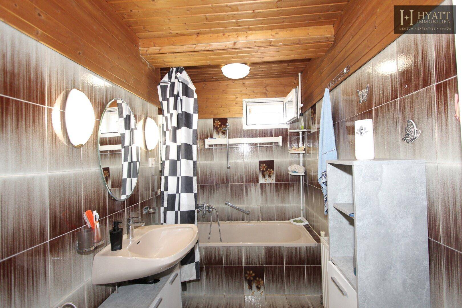 Badezimmer mit braunen Fliesen, Badewanne und Holzdecke im Stil der 80er Jahre.