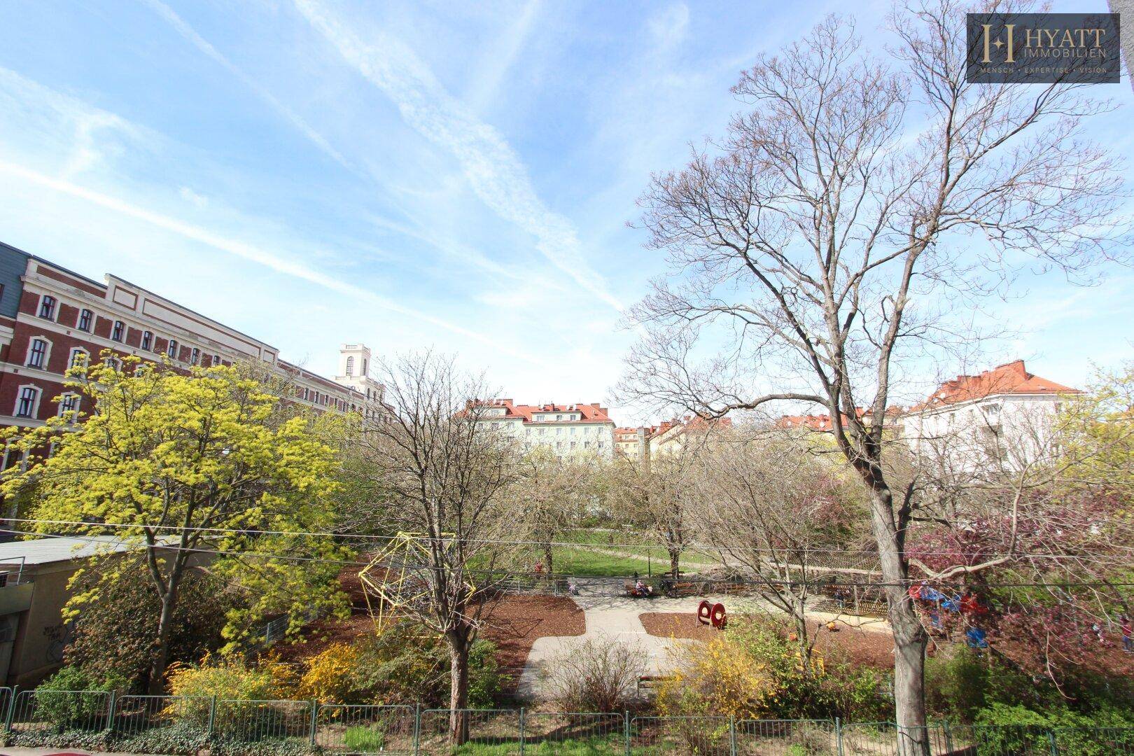 Weitläufiger Blick auf einen grünen Park mit Spielplatz und umliegenden Gebäuden.