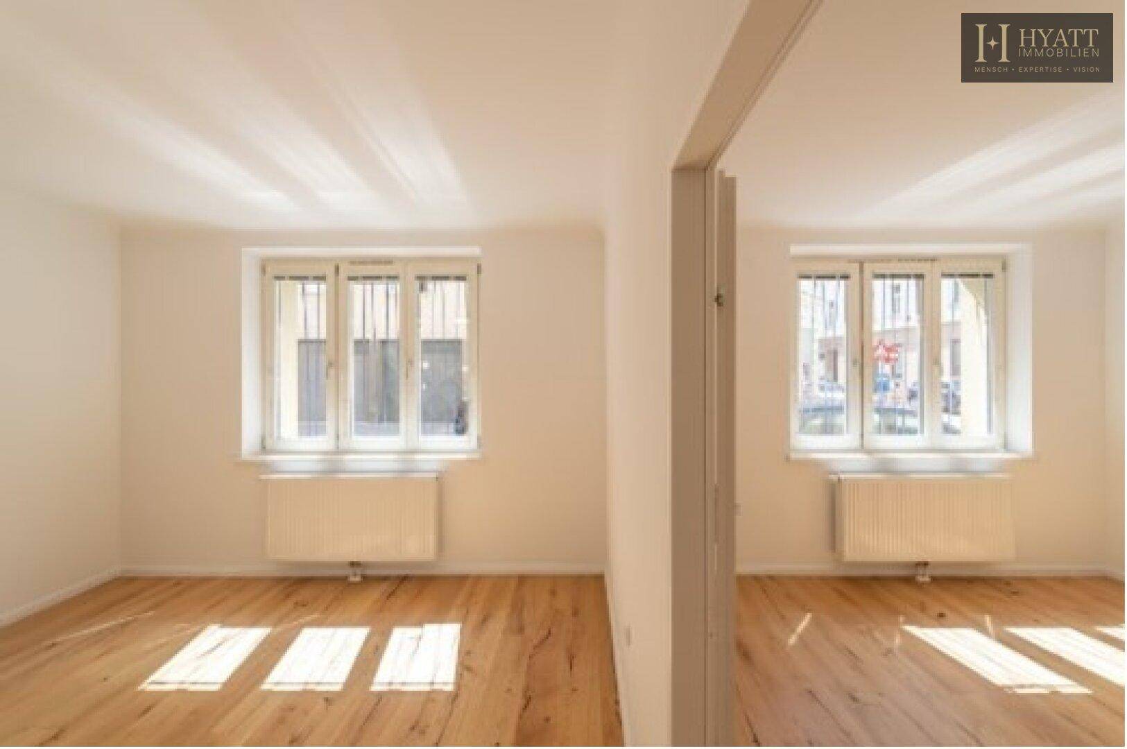 Heller, leerer Raum mit Holzboden und zwei Fenstern, die viel natürliches Licht hereinlassen.