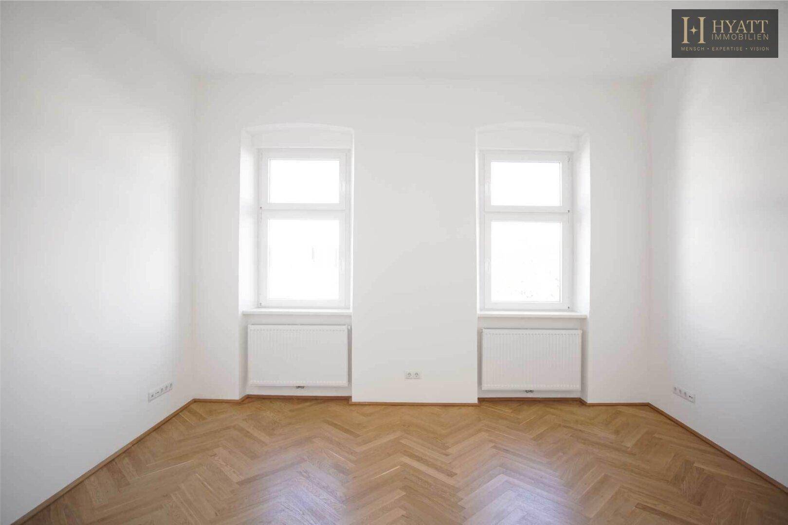Unmöblierter Raum mit Fischgrätparkett und zwei hohen Fenstern, ideal für individuelle Gestaltung.