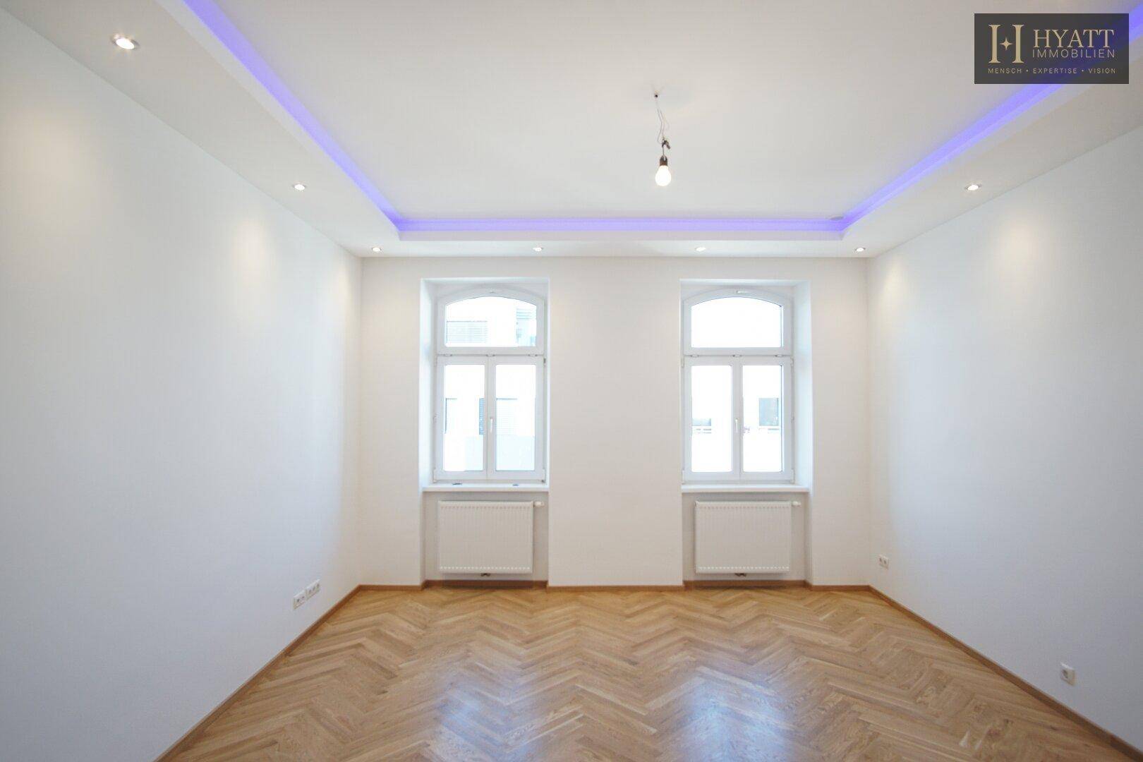 Helles, leeres Zimmer mit Fischgrätparkett, hohen Decken und indirekter Deckenbeleuchtung.