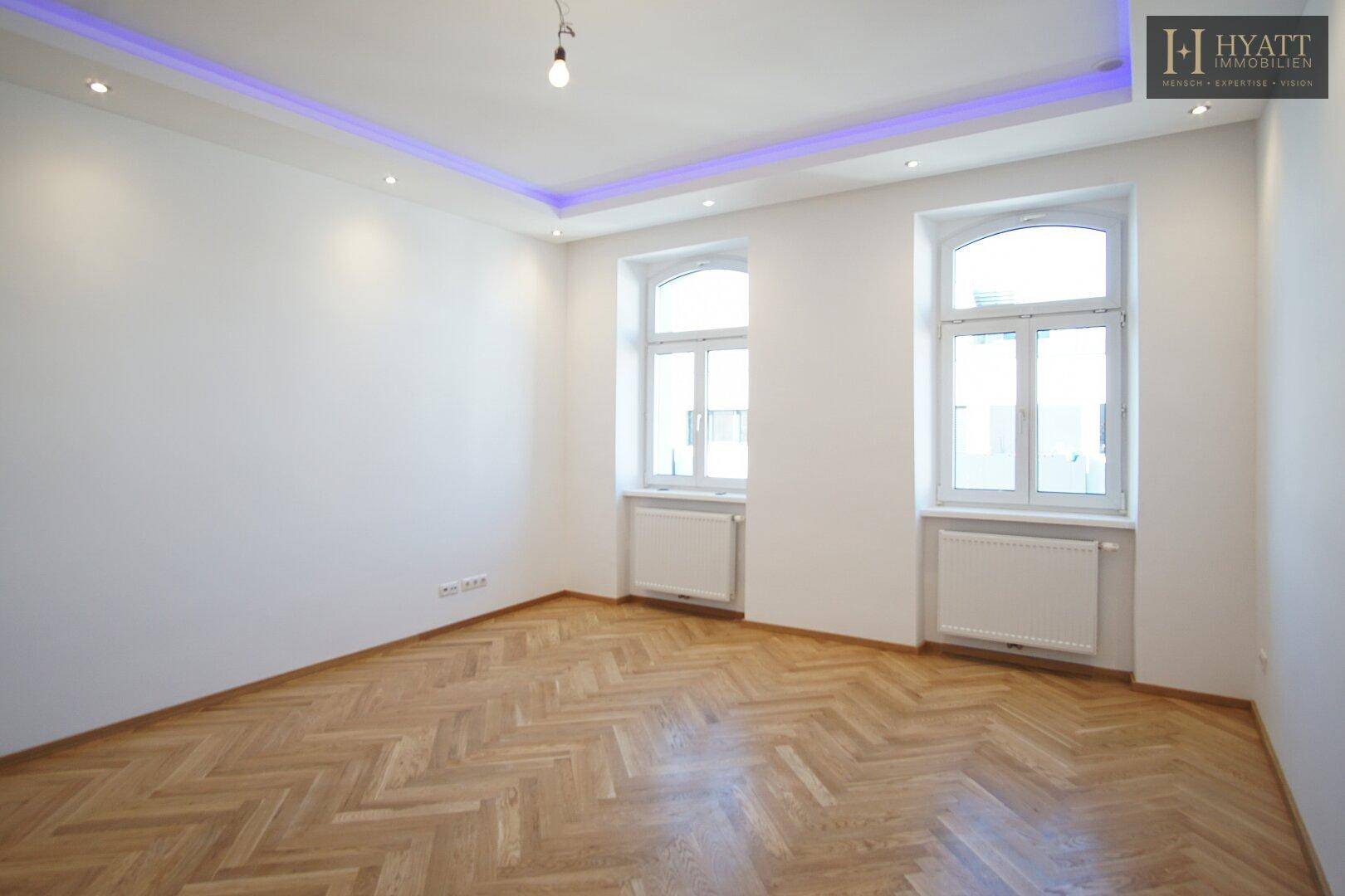 Leeres Zimmer mit Fischgrätparkett, hohen Decken und indirekter Deckenbeleuchtung.