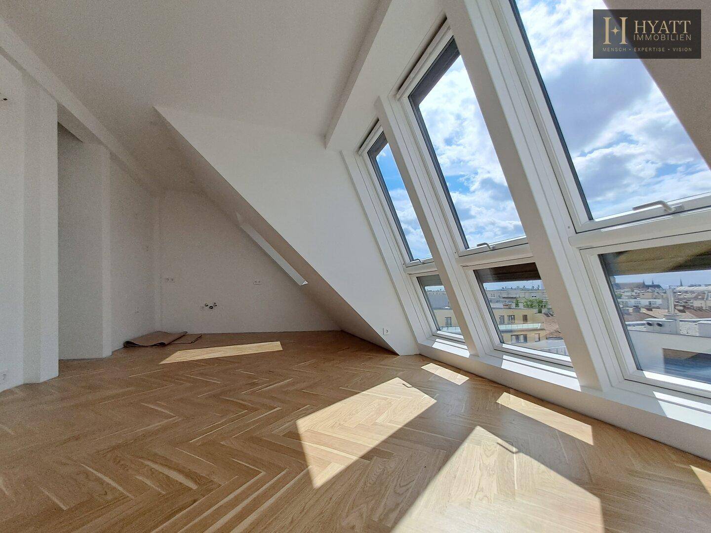 Heller Raum mit Fischgrätparkett und großen, schrägen Fenstern, die einen weiten Blick über die Stadt bieten.