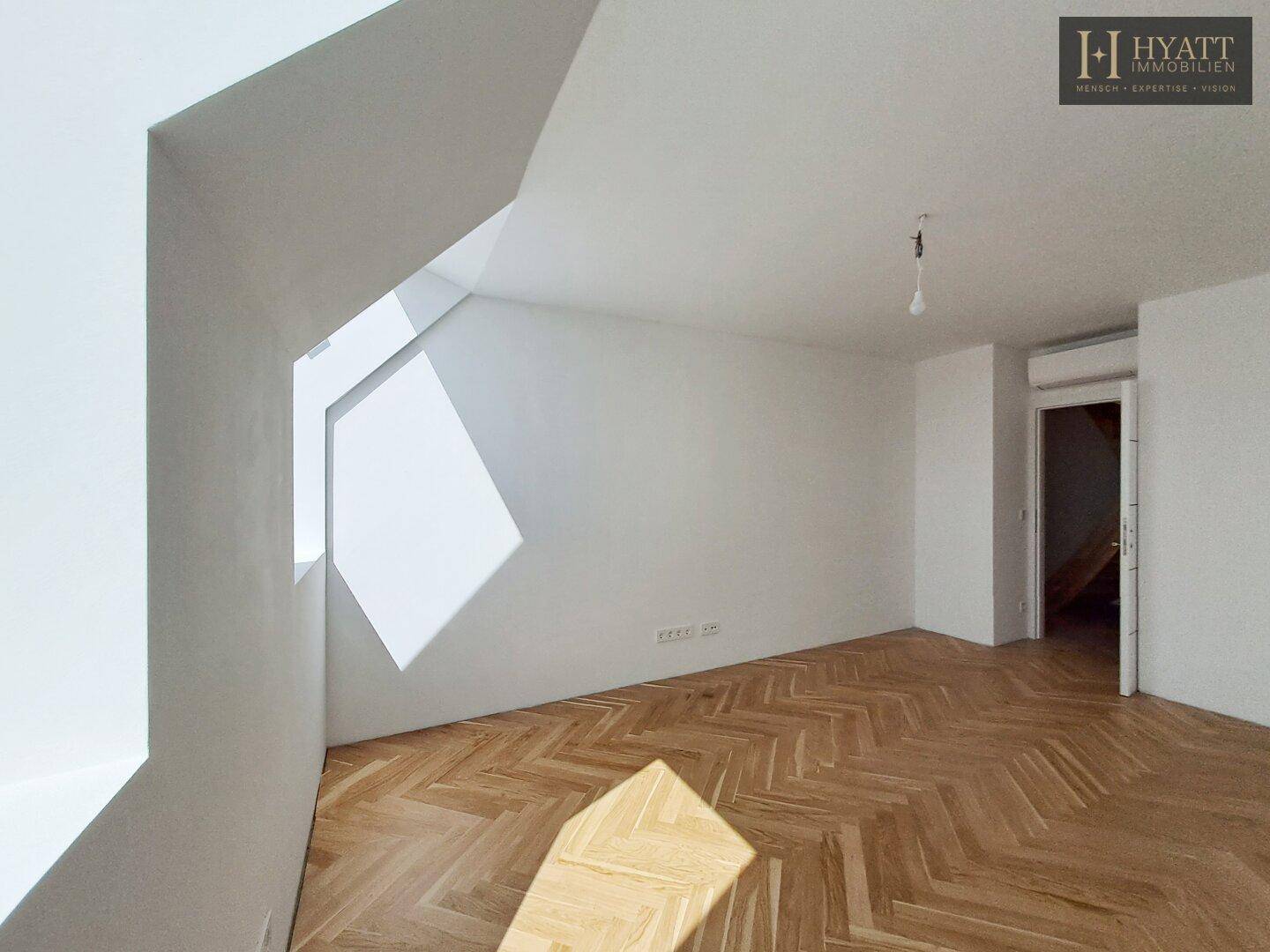Leeres Schlafzimmer mit Fischgrätparkett, weißen Wänden und einem schrägen Fenster, das viel Licht spendet.