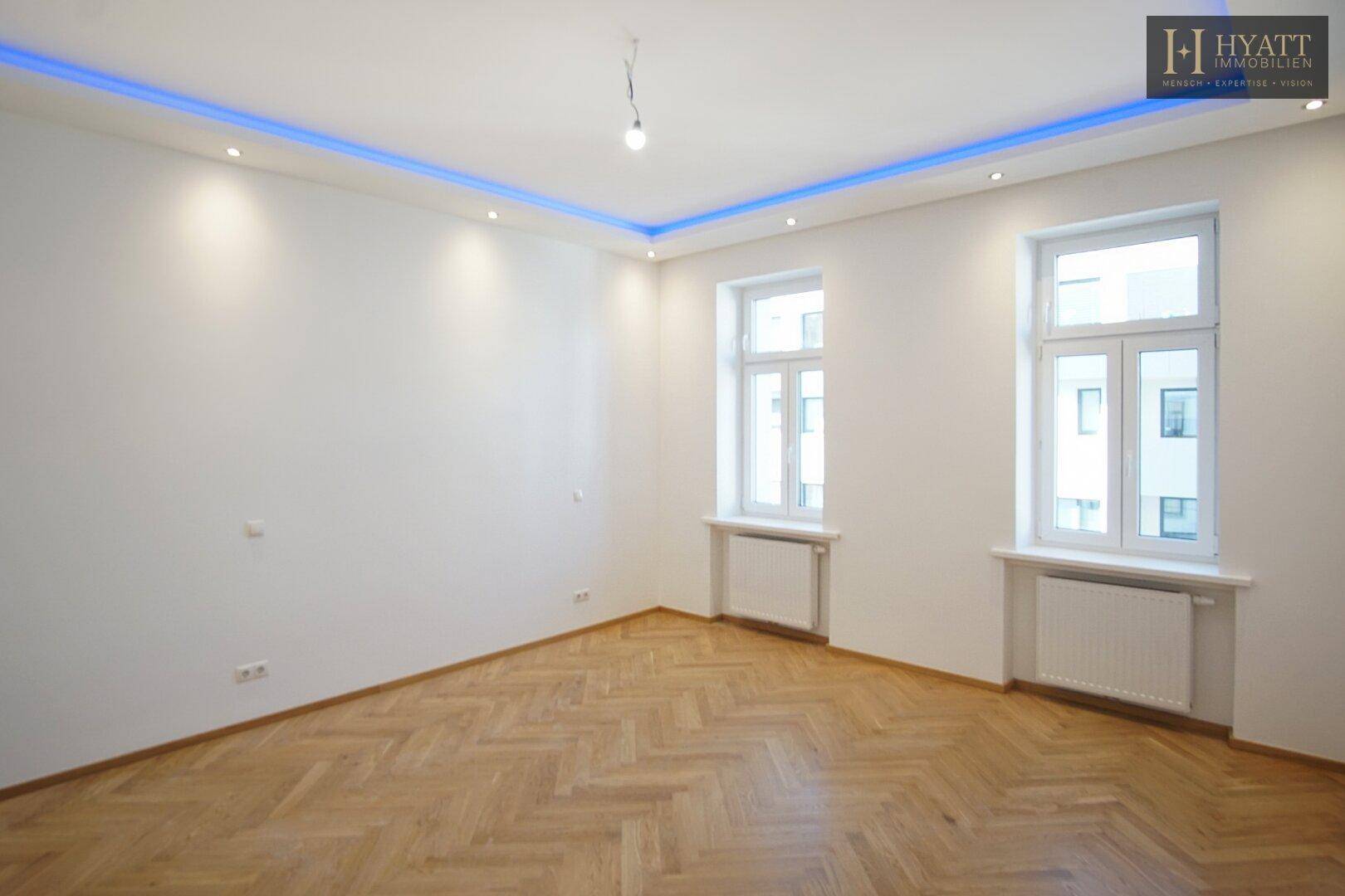 Leerer Raum mit Fischgrätparkett, großen Fenstern und moderner Deckenbeleuchtung.