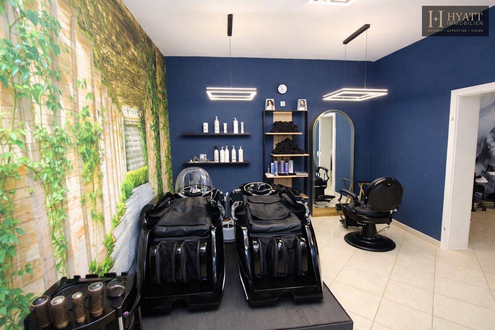 Geräumiger Schönheitssalon mit zwei Spa-Liegen, einem Friseurstuhl und einer grünen Wandtapete mit Pflanzenmotiv.