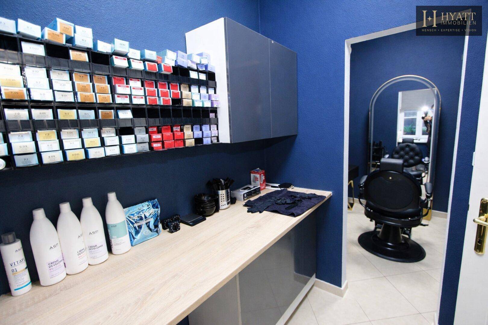 Arbeitsbereich für Haarfärbemittel mit zahlreichen Produkten, Ablageflächen und einem Friseurstuhl im Hintergrund.