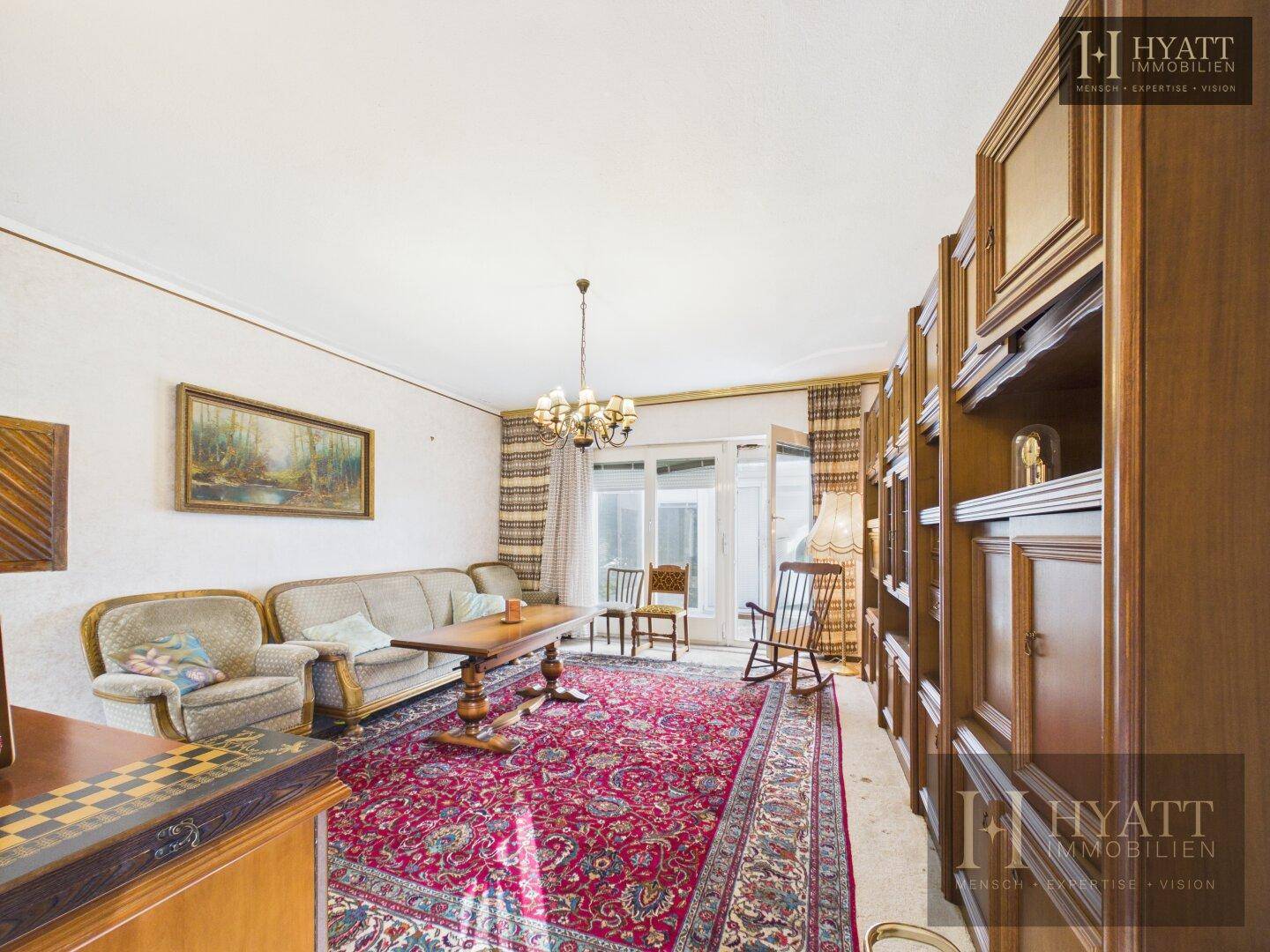 Gemütliches Wohnzimmer mit traditionellen Möbeln, großem Teppich und Zugang zum Balkon.