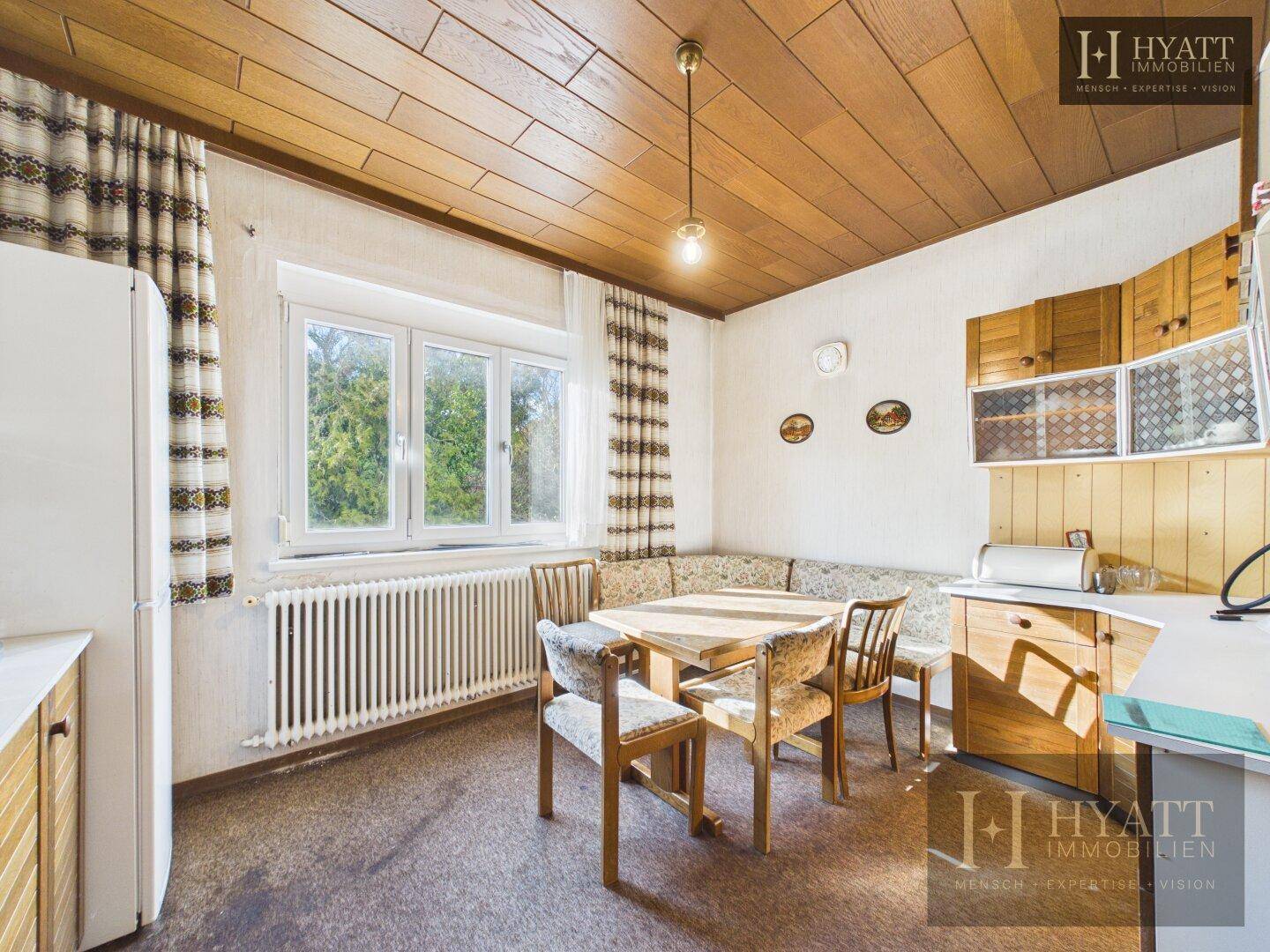 Gemütliche Wohnküche mit Esstisch, Holzdecke und großem Fenster, das viel Tageslicht hereinlässt.