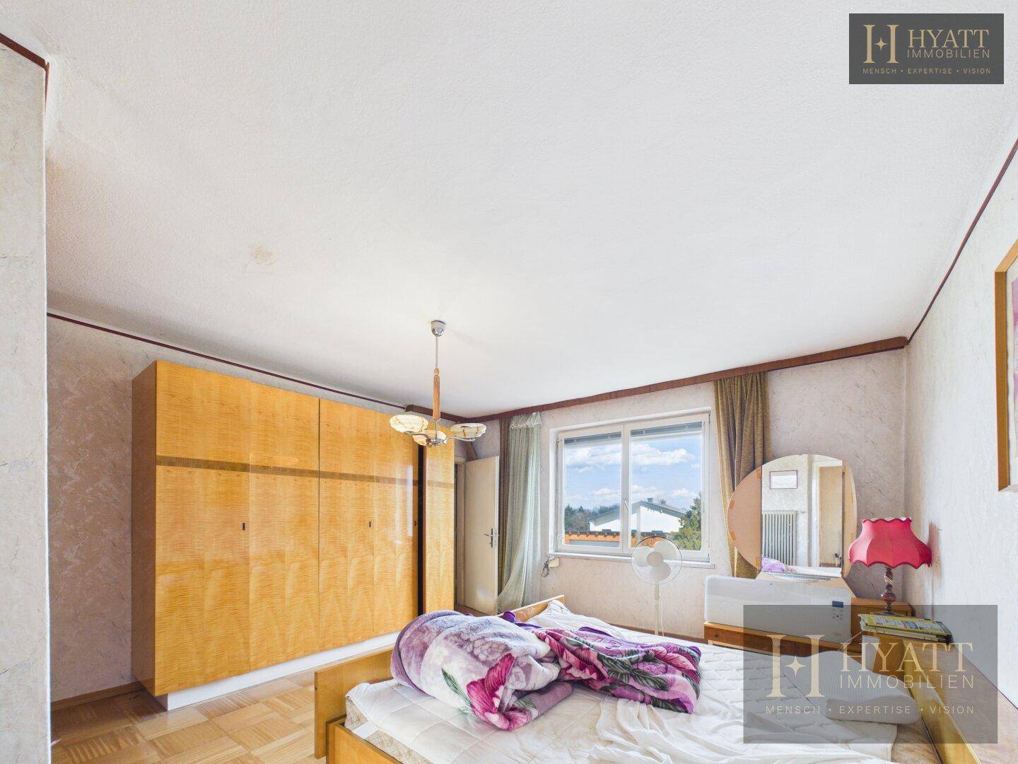 Schlafzimmer mit Bett, großem Holzschrank und Fenster, das viel Tageslicht hereinlässt.