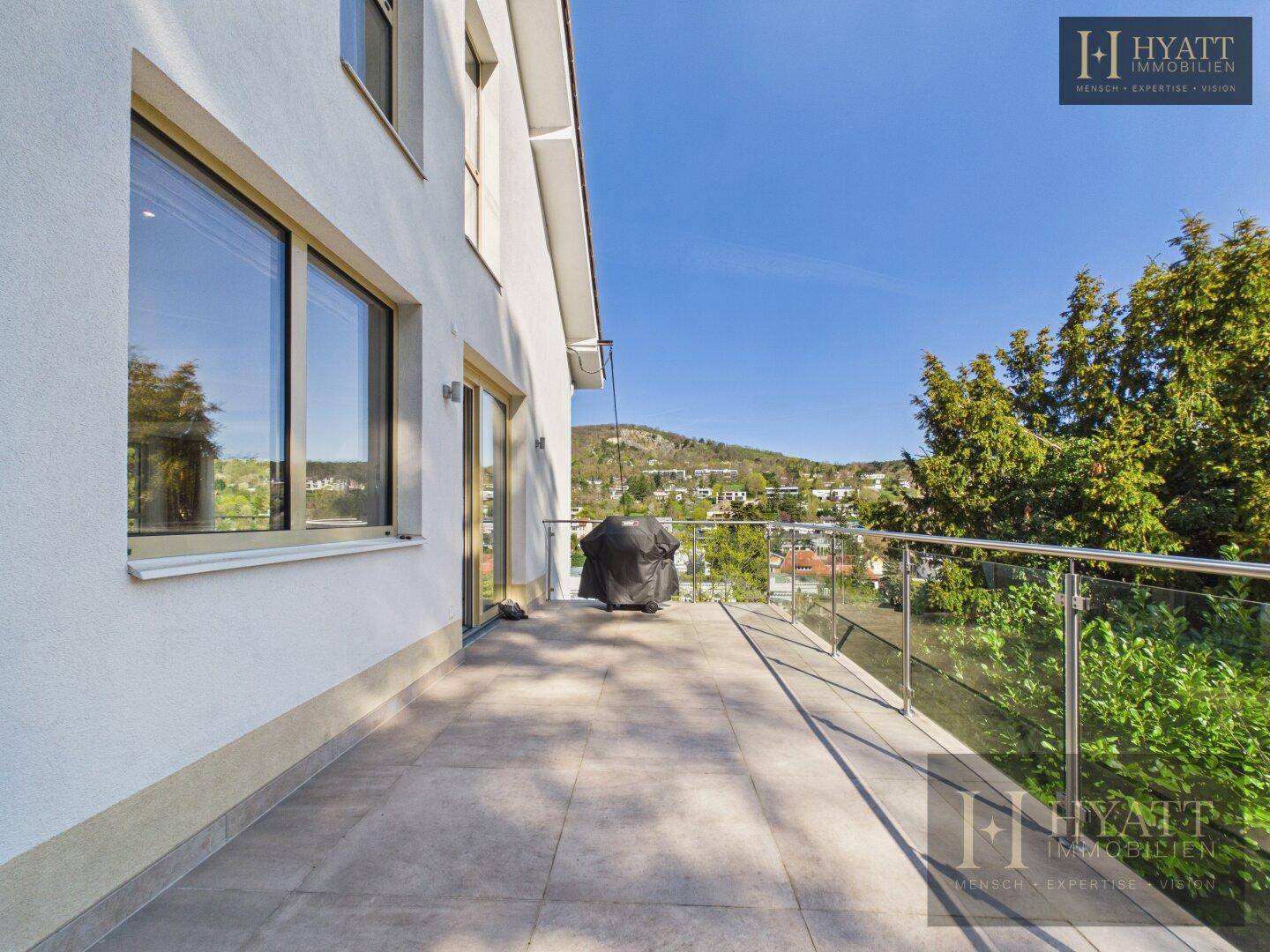 Geräumige Terrasse mit Fliesenboden, Glasgeländer und Grill, bietet einen weiten Blick ins Grüne.