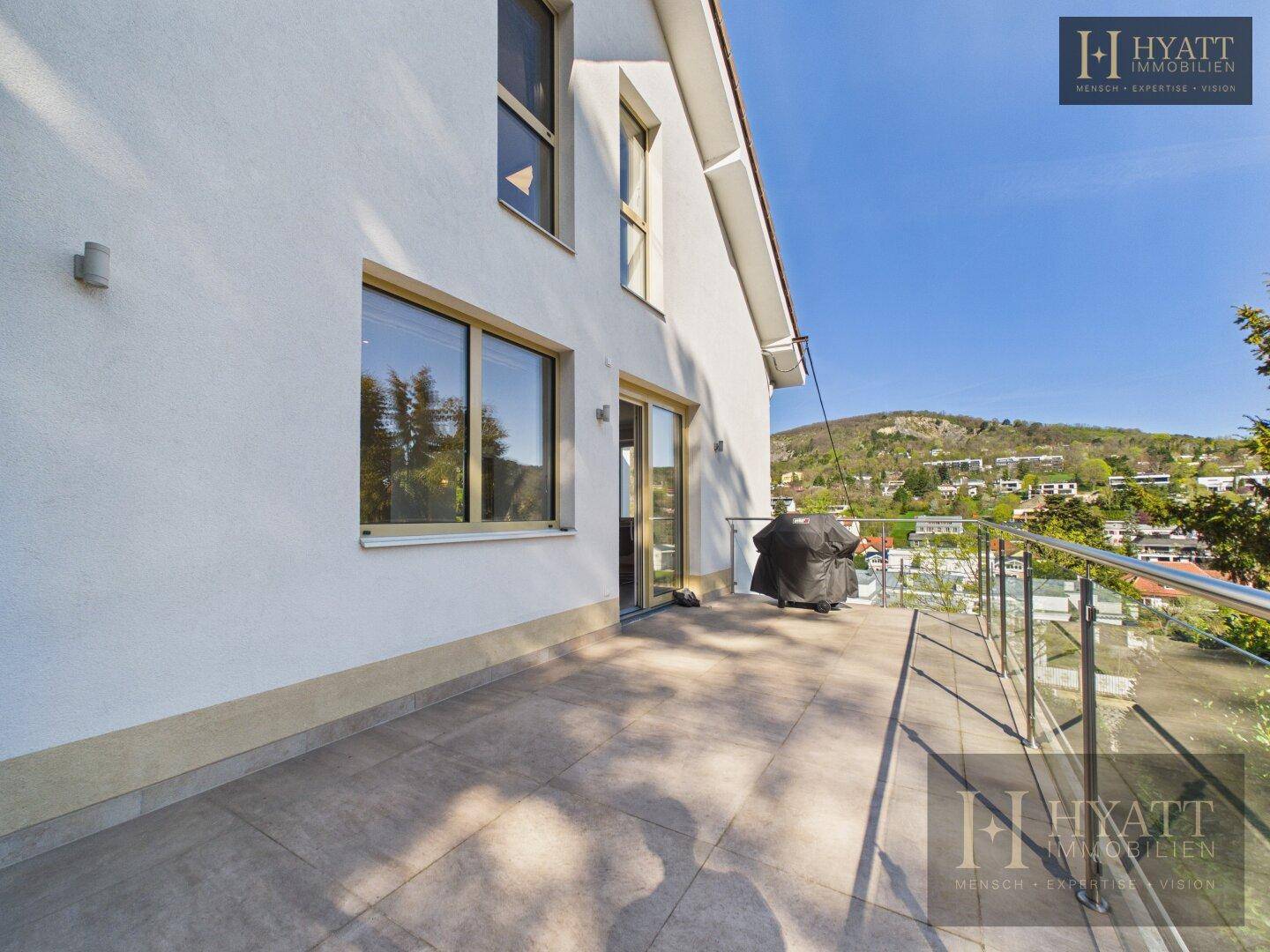 Sonniger Terrassenbereich mit modernen Fliesen, Glasgeländer und Blick auf die hügelige Landschaft.