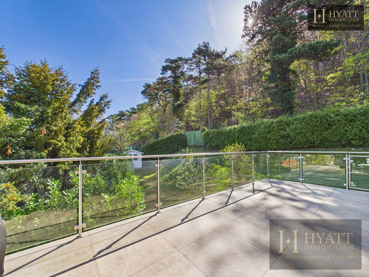Große Terrasse mit Glasgeländer und direktem Zugang zu einem gepflegten Gartenbereich mit Bäumen.