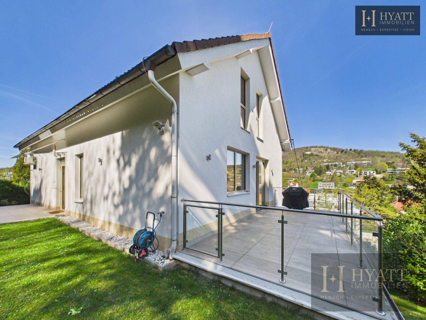 Geräumige Terrasse mit Glasgeländer am Haus, umgeben von Grünflächen.