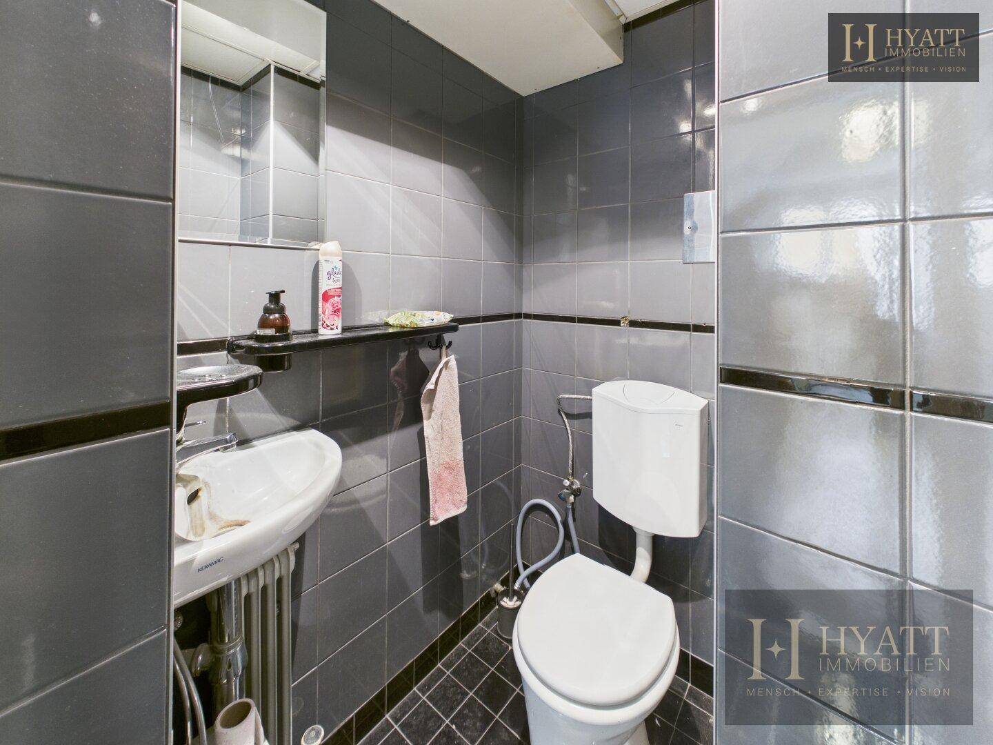 Kleines Badezimmer mit grauen Fliesen, Waschbecken und Toilette.