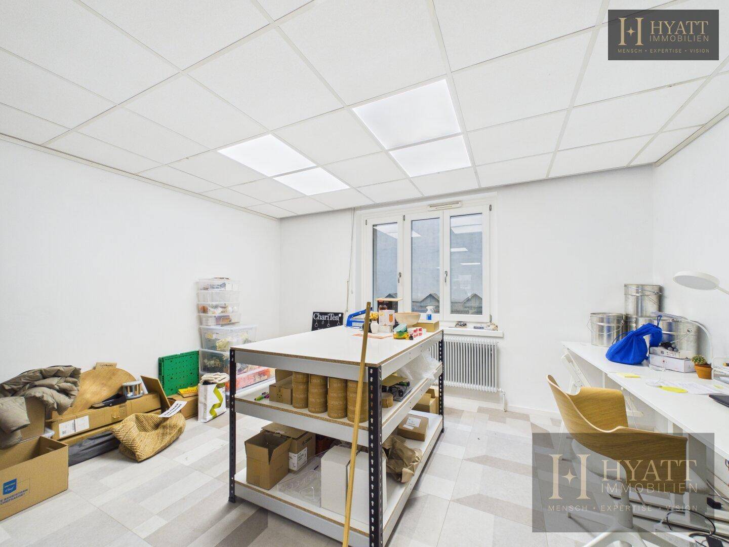 Heller Büroraum mit großem Arbeitstisch, Fenster und abgehängter Decke.