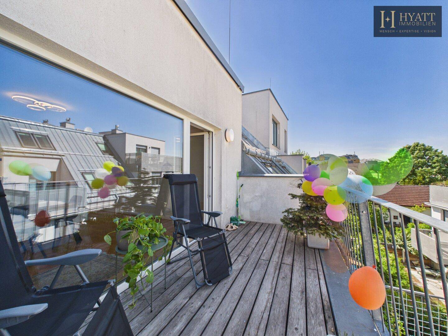 Sonniger Balkon mit Holzdeck, Pflanzen und Blick über die Dächer der Stadt.