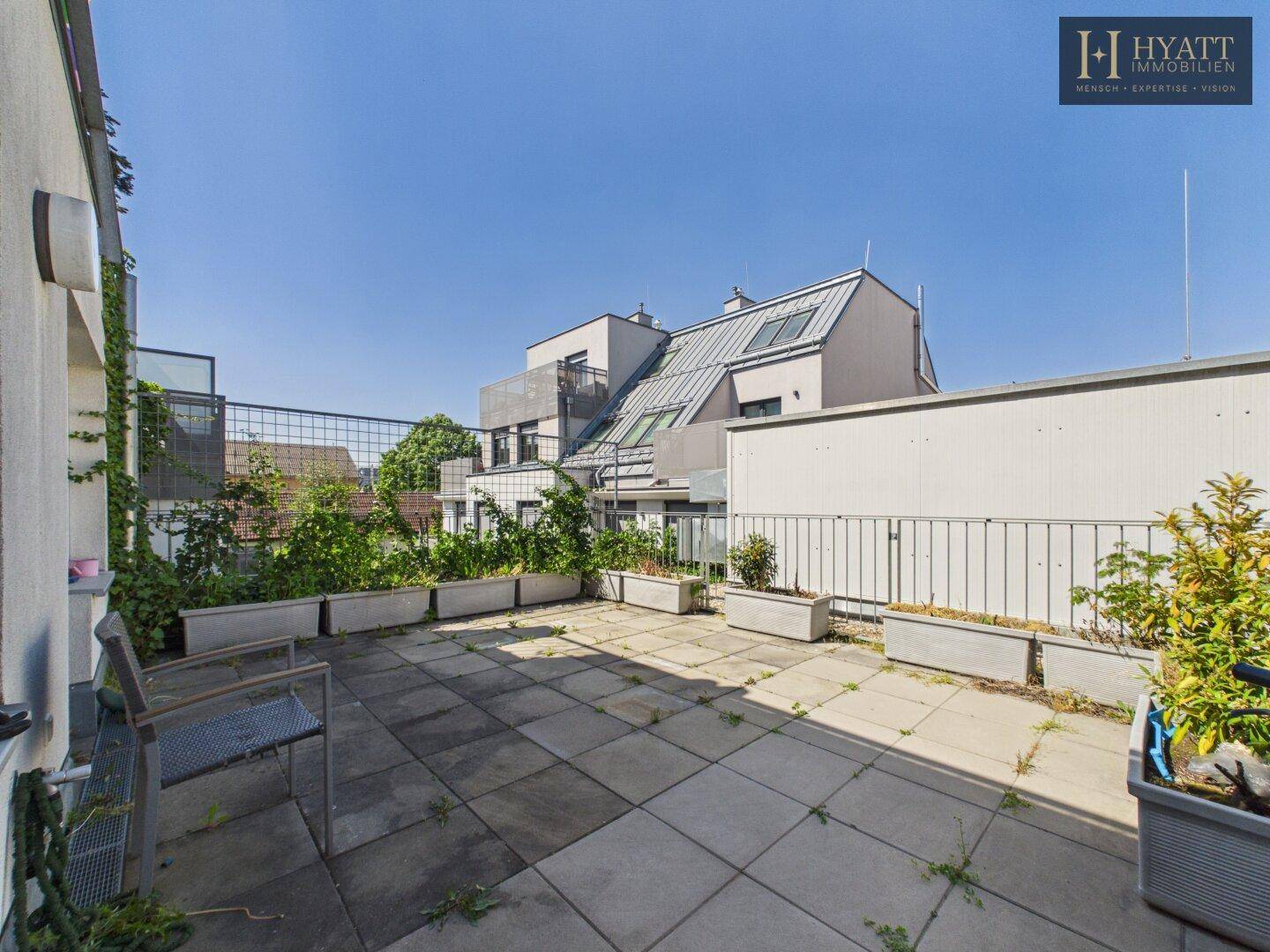 Weitläufige Dachterrasse mit Steinplatten, Pflanzen und Blick auf die umliegenden Gebäude.