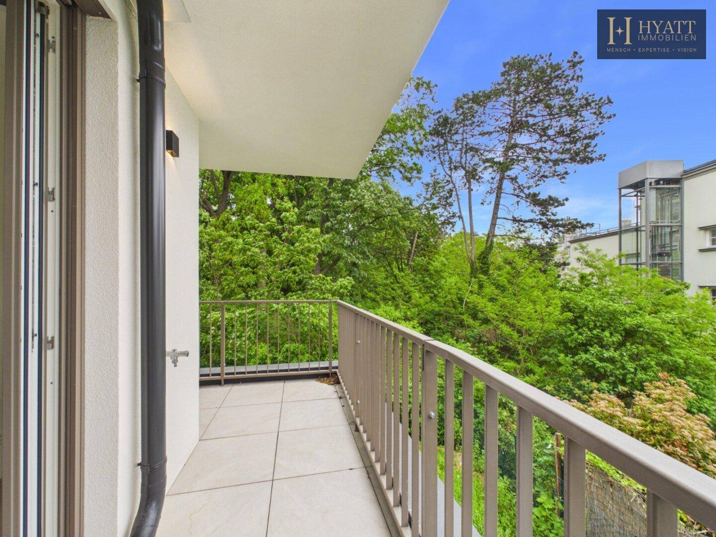 Geräumiger Balkon mit Fliesenboden und Blick auf grüne Bäume und umliegende Gebäude.