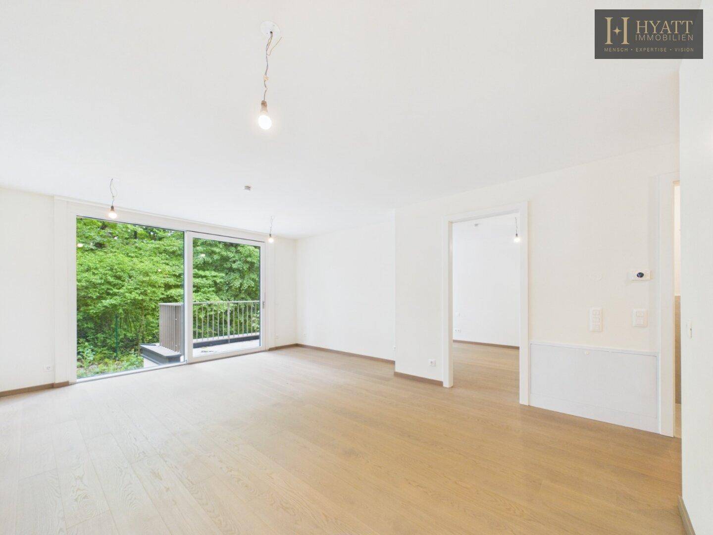 Geräumiges, leeres Wohnzimmer mit hellem Holzboden und großen Fenstern mit Blick ins Grüne.